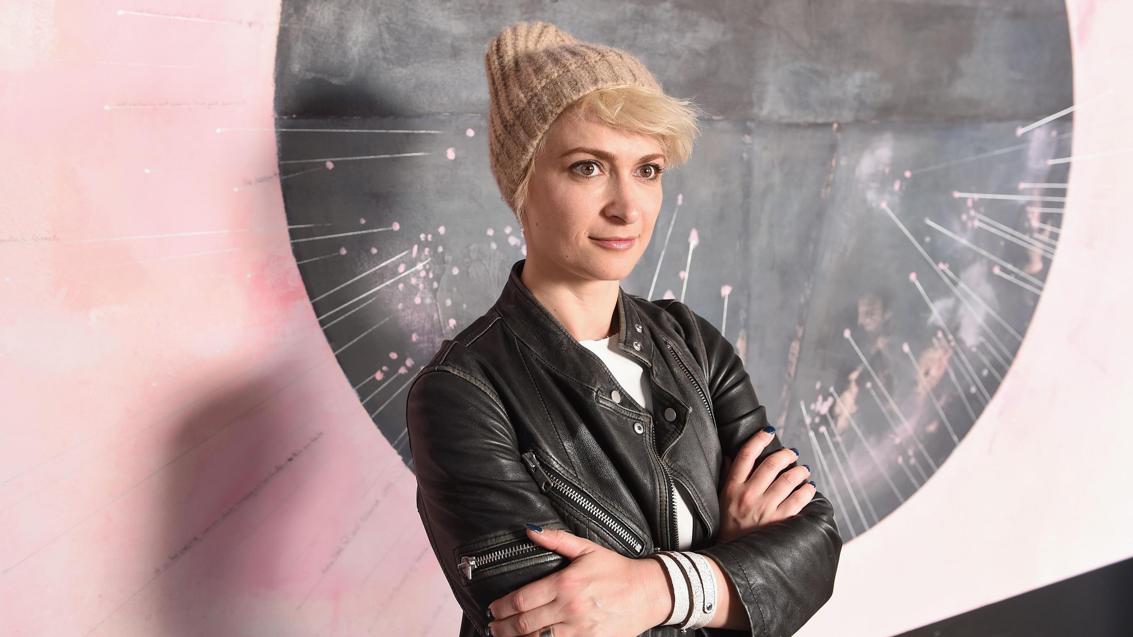 Hutchins is wearing a woolly hat and leather jacket and stands with her arms crossed