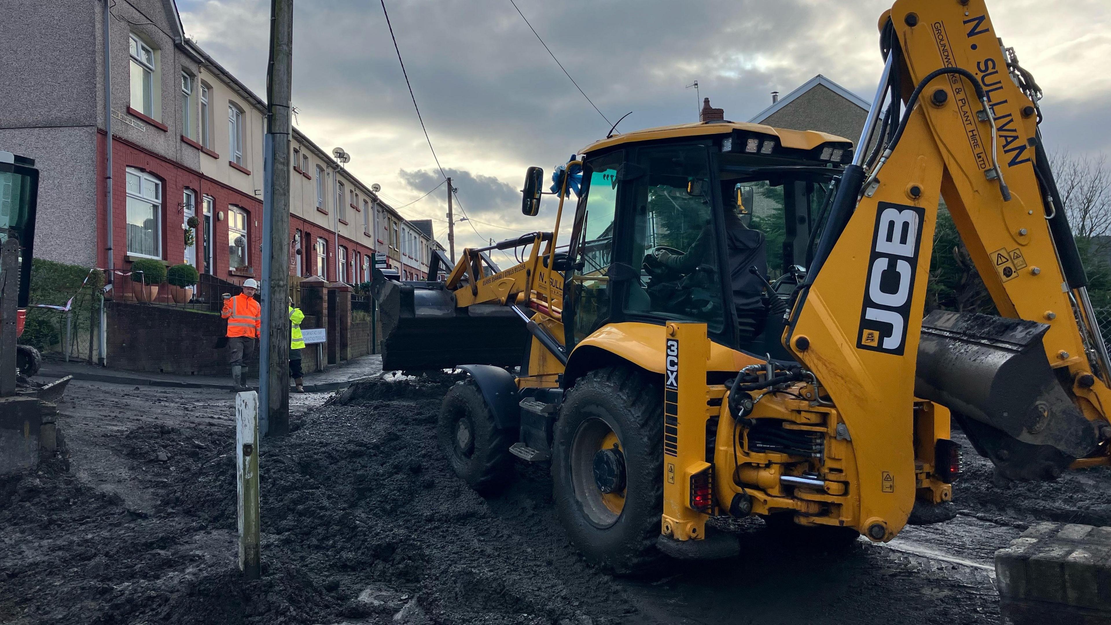 A large yellow digger moving slurry piled onto a street in Cwmtillery. Workmen in high viz jackets and hard hats are on the pavement across from the digger. 