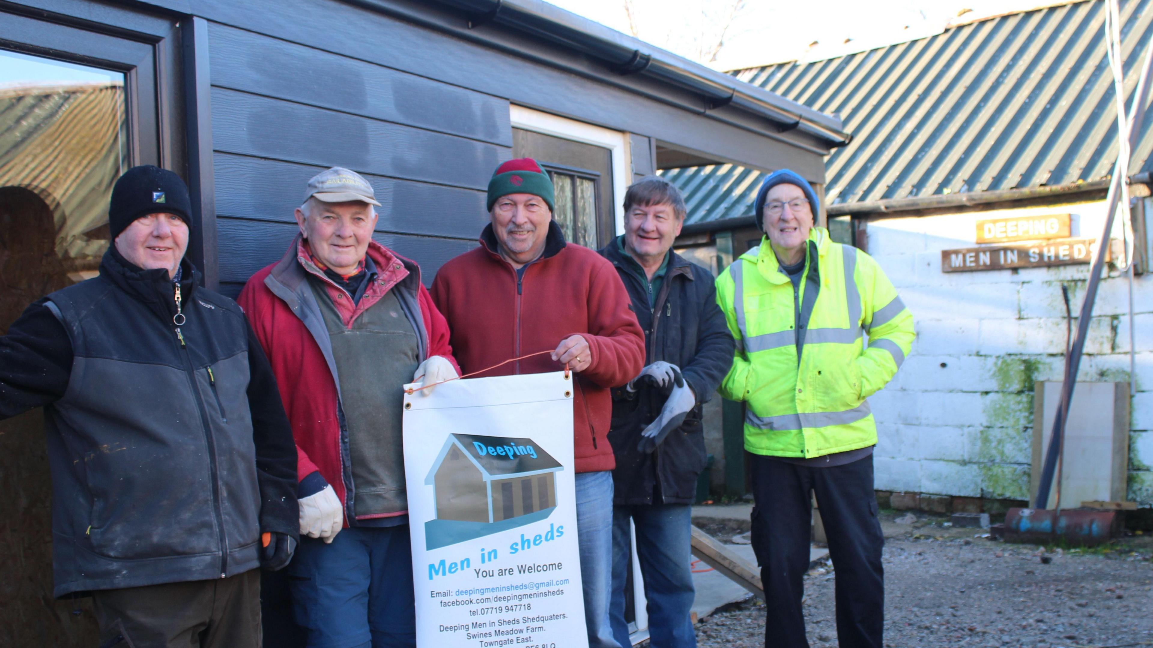 Members of the Deepings Men in Sheds group standing in from of a grey-coloured building. They are holding a placard which says: "You are welcome."