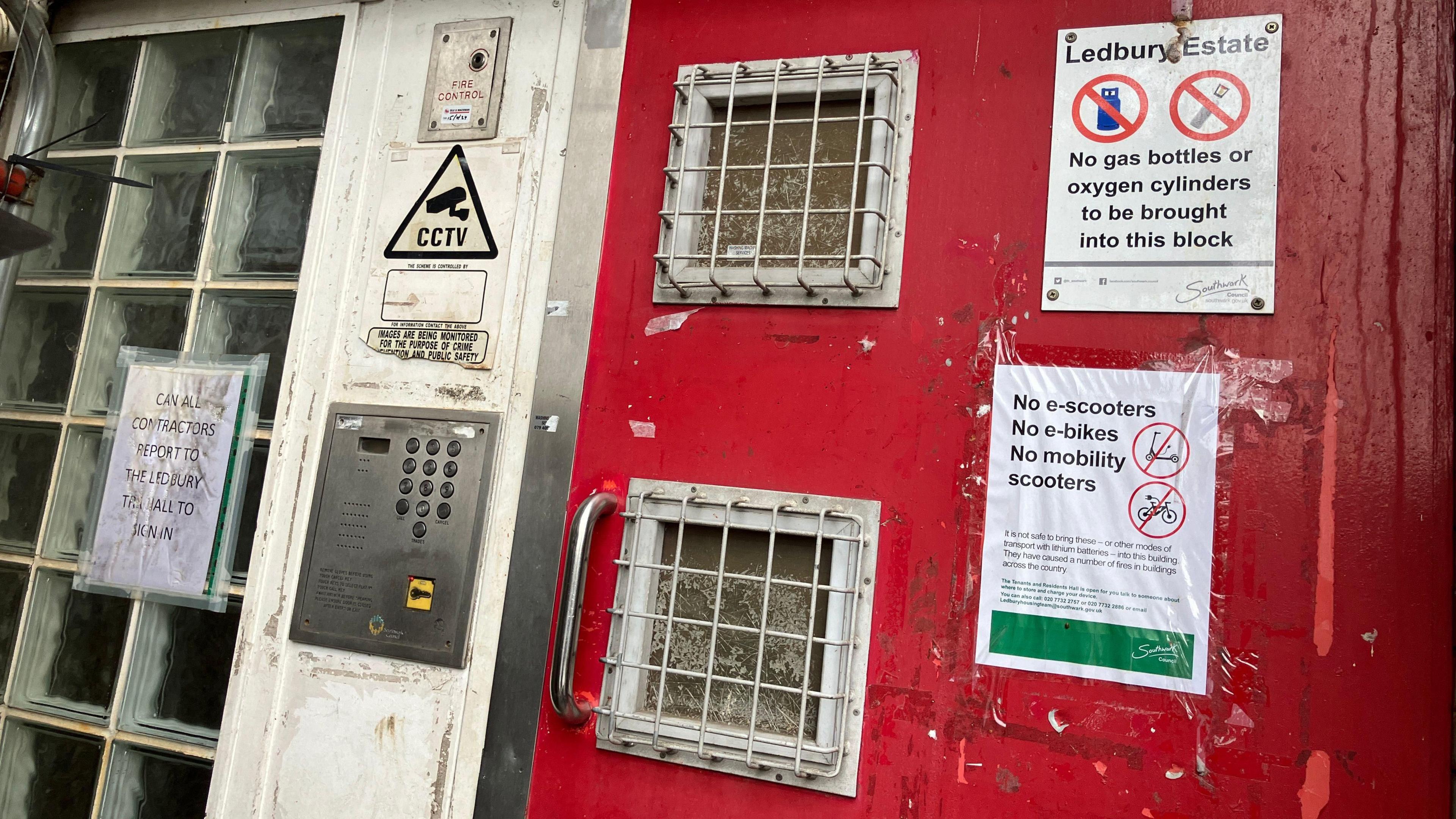 The entrance to one of blocks on the Ledbury Estate - a bright red steel door with small porthole windows covered in metal gratings and multiple white warning signs saying 'no e-scooters, no e-bikes' and 'no gas bottles'