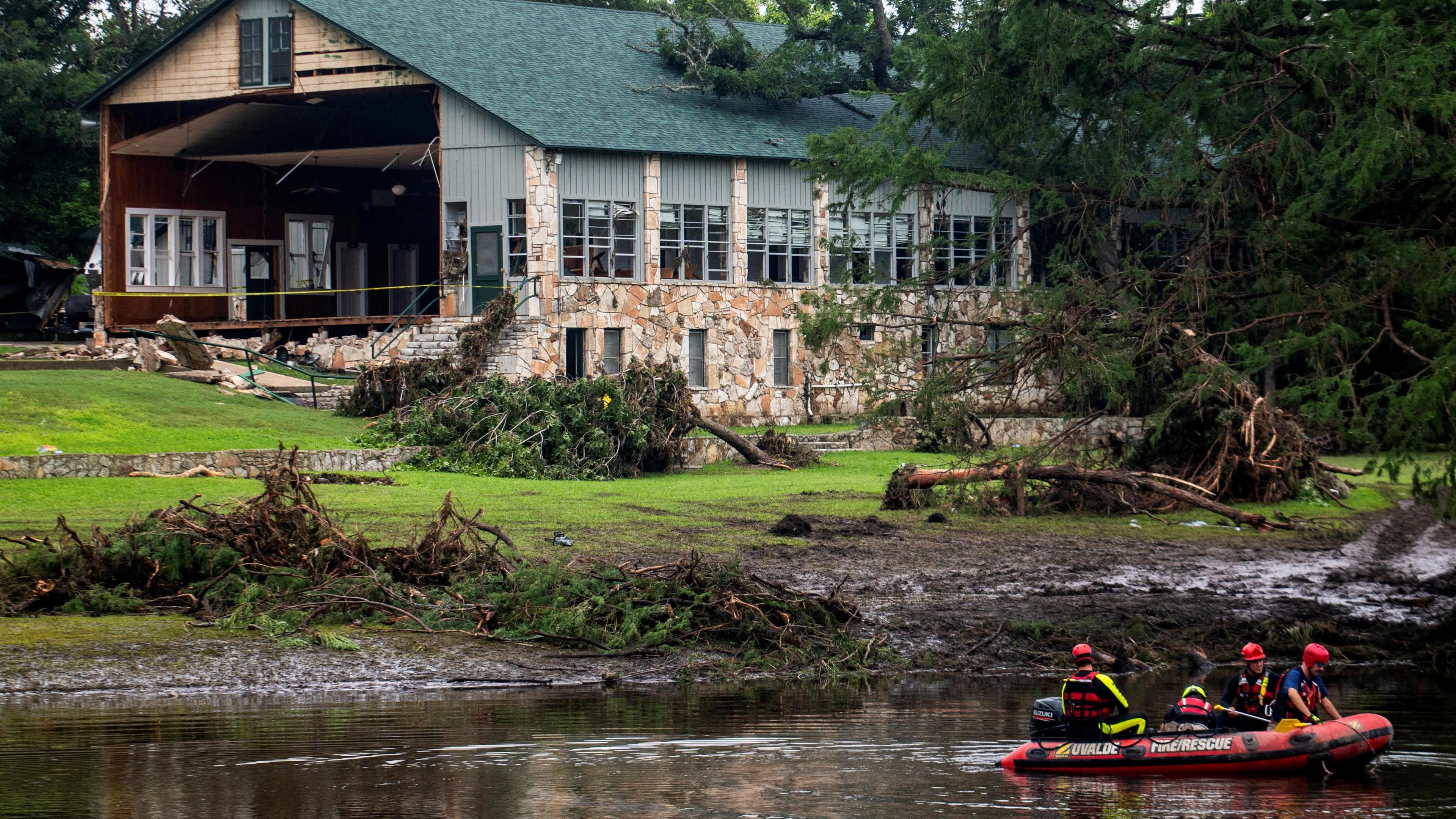 People in a boat row down a river with a damaged building in the background and trees strewn over grass