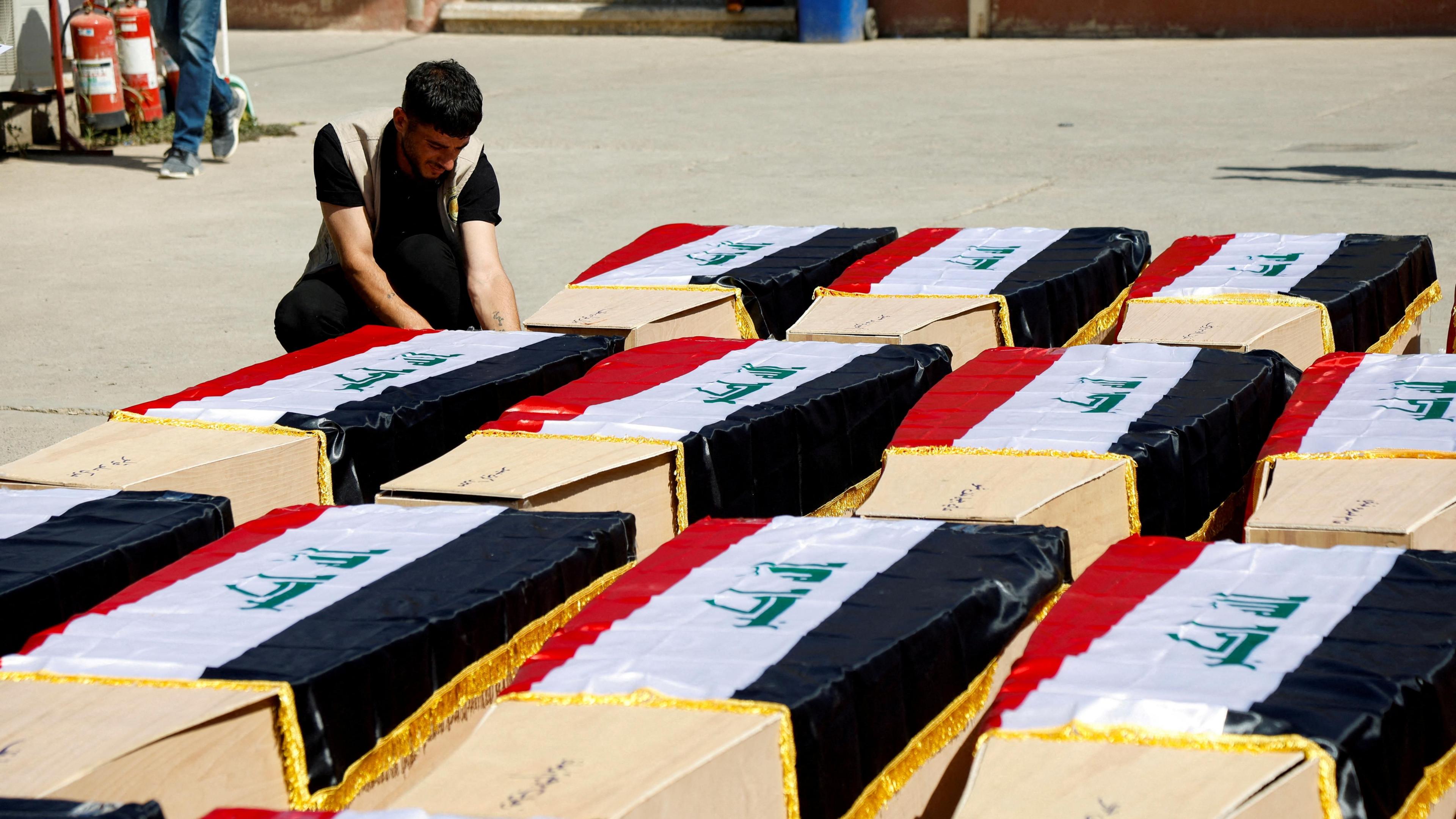 A man sits next to rows of coffins containing the remains of Yazidis killed by the Islamic State group in 2014, following the exhumation of a mass grave in Mosul, Iraq (20 June 2023)