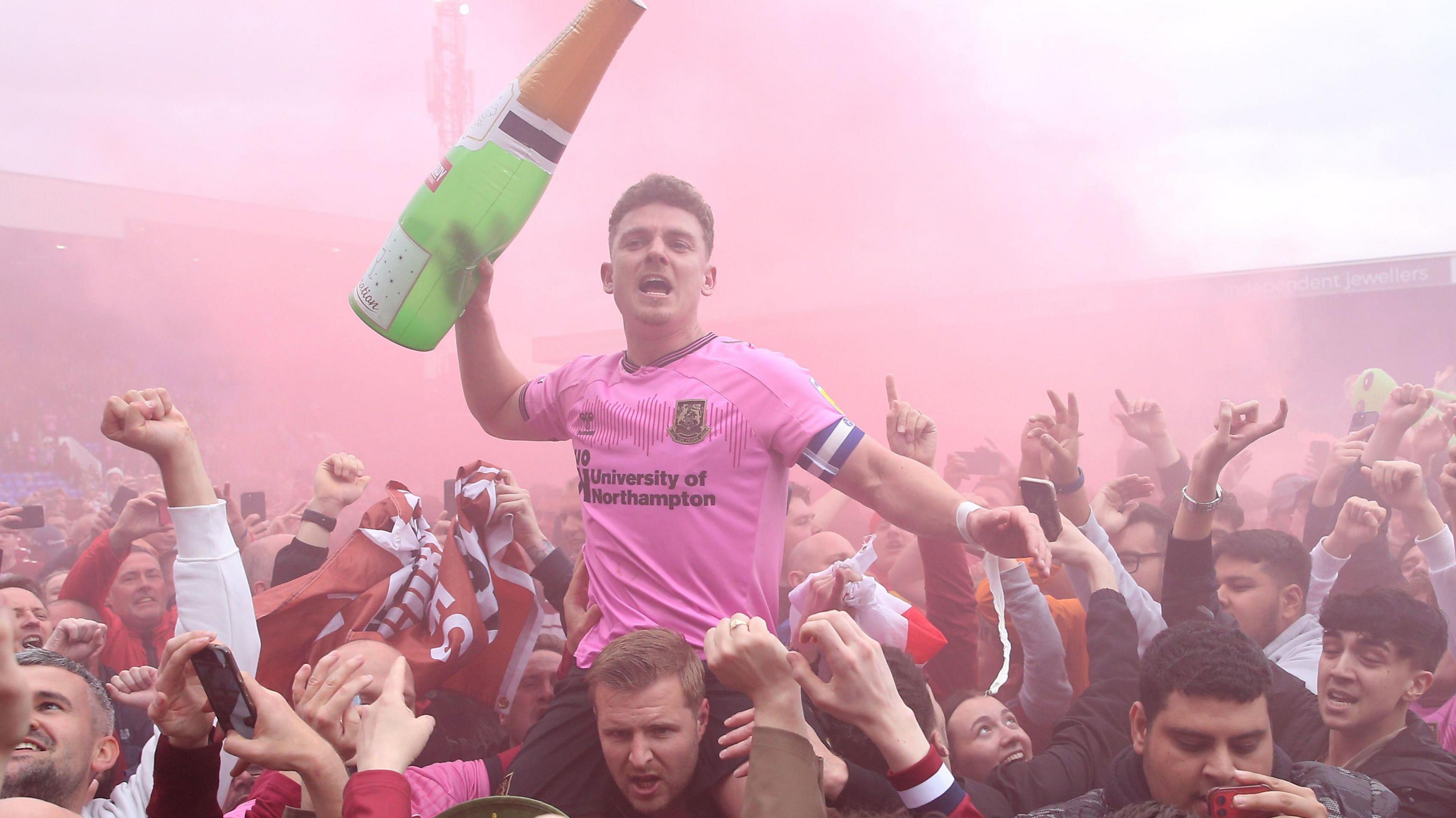 Sam Hoskins in a pink Northampton Town shirt on the shoulders of a fan with an inflatable champagne bottle among a sea of supporters and smoke from a flare.