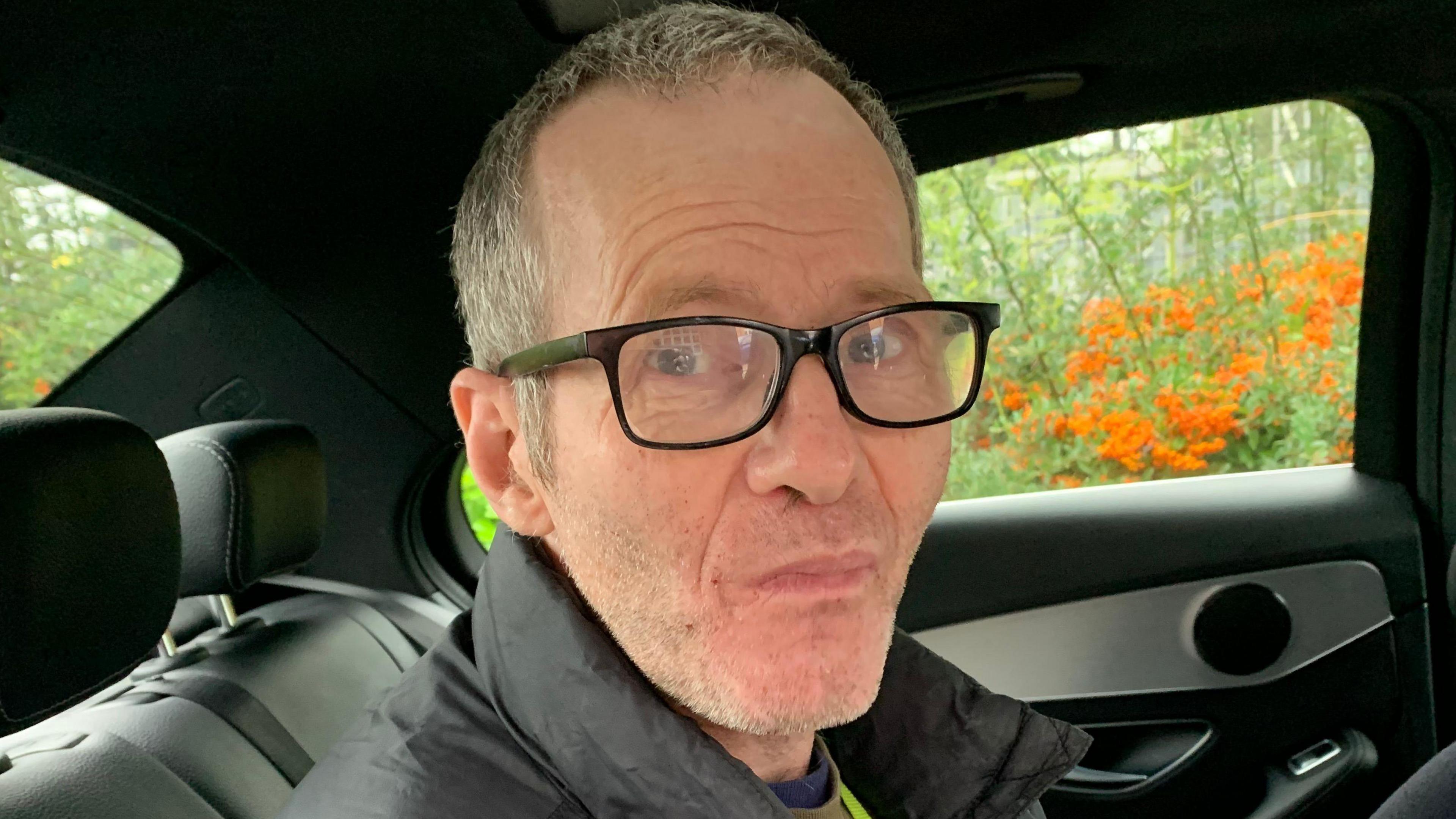 Close-up of Alan Jones, who has short hair and stubble, looks into the camera. He is wearing glasses and sitting in the back of a car.