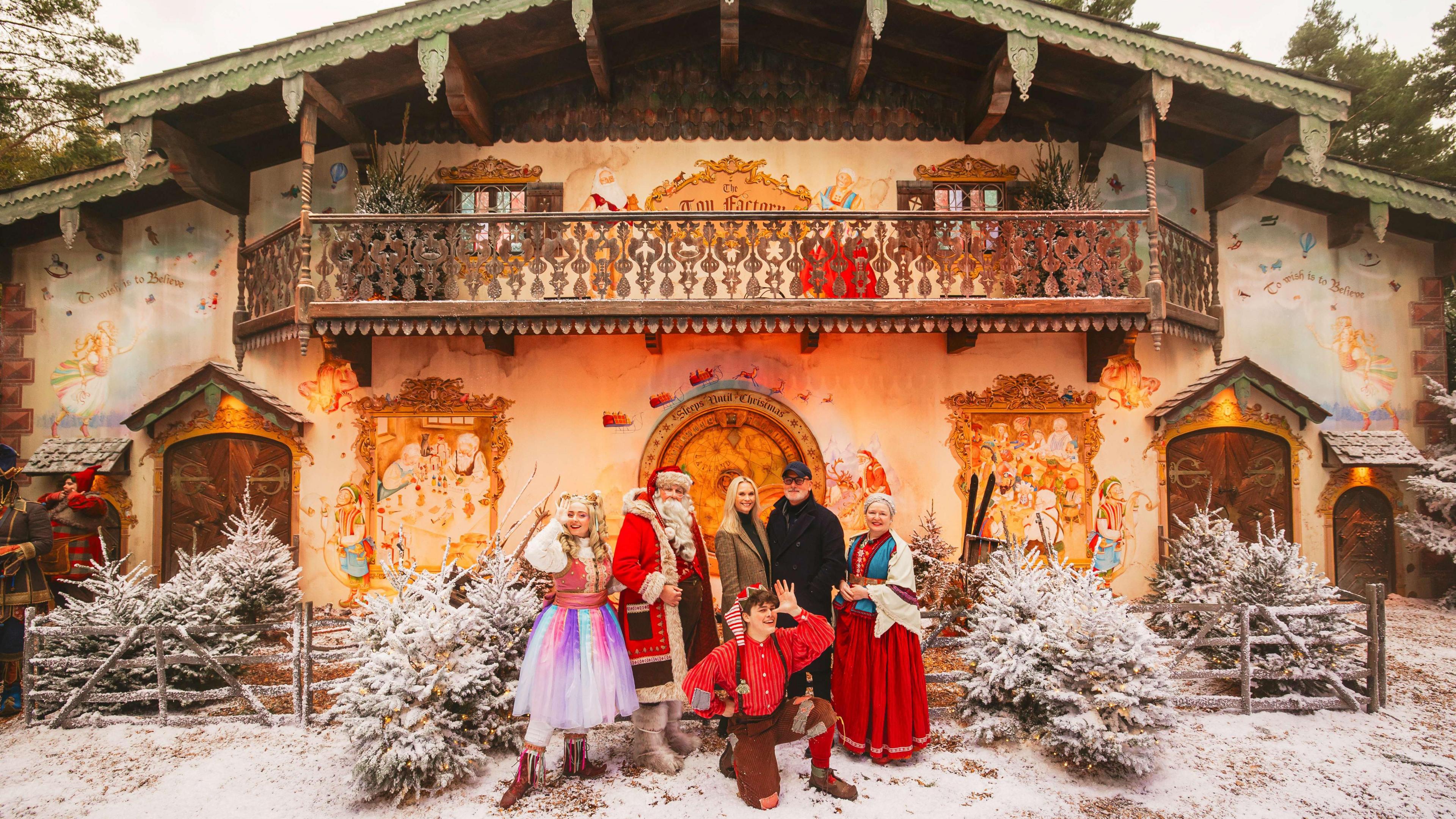 Cast members at Lapland UK stand in front of a house surrounded by Christmas trees, wearing Christmas outfits. snow on the ground. and the building has a balcony and a sign saying Toy Factory. Father Christmas is in the crowd with elves and members of the public. 