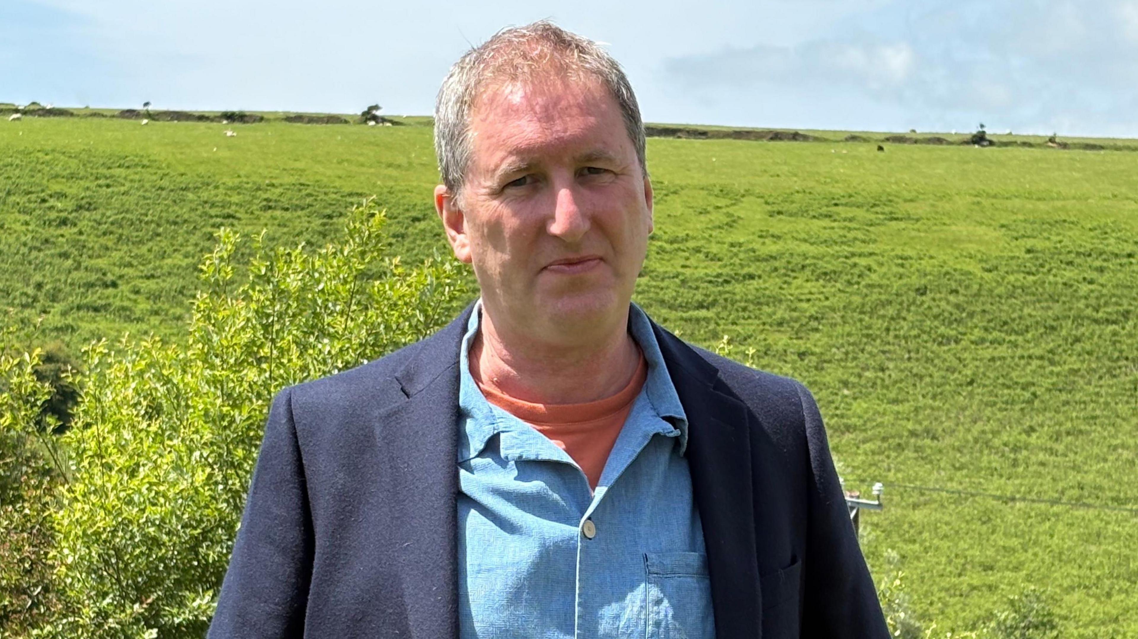 A man with grey hair wearing an orange t-shirt, blue shirt, and dark blue jacket standing in front of a green hill with sheep grazing on it.