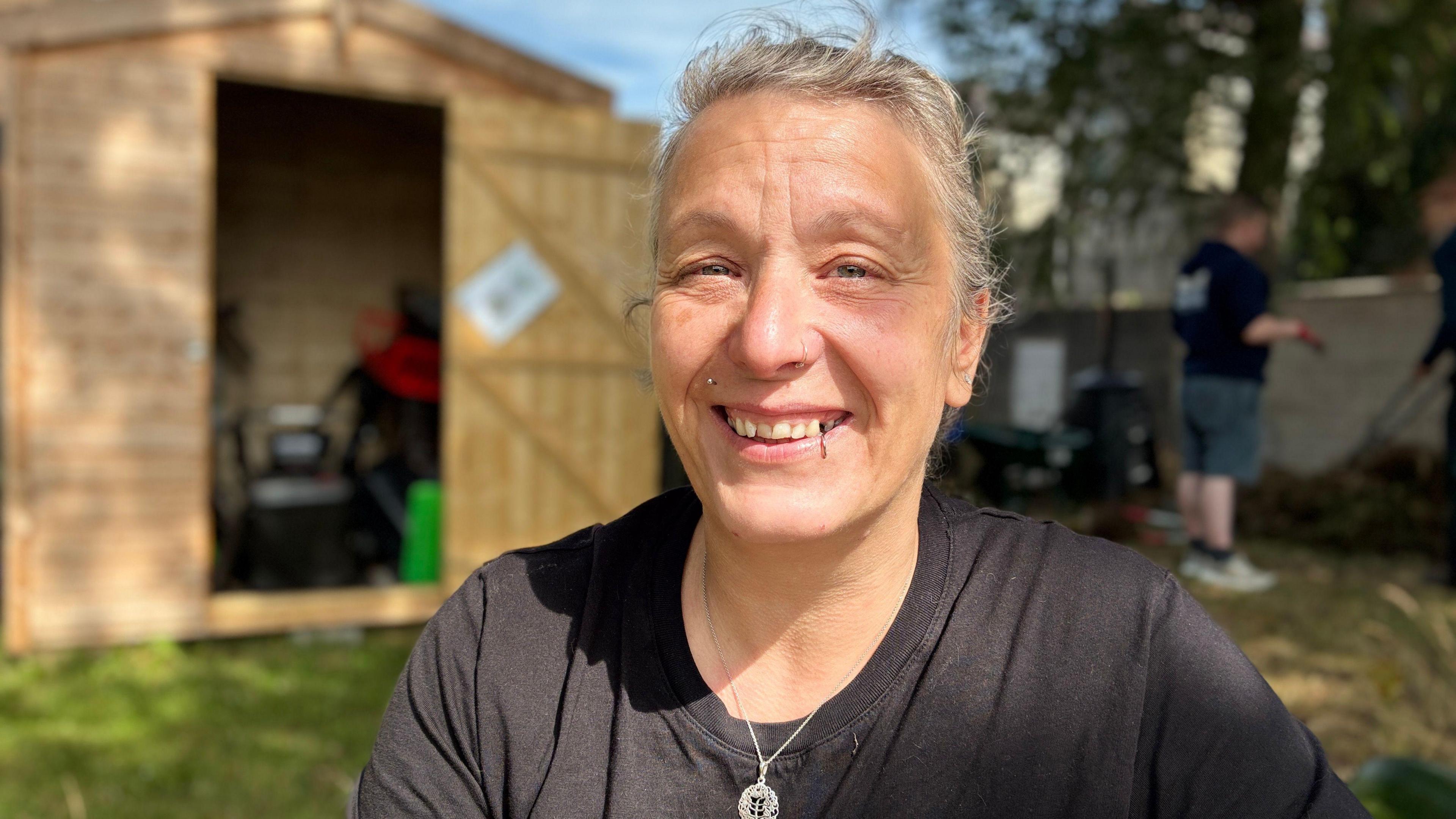 A smiling woman sits outdoors in front of a wooden garden shed with its door open, revealing tools and equipment inside. She wears a black T-shirt and a silver pendant necklace, her hair loosely tied back. The sunlight highlights her face, giving the scene a warm, friendly atmosphere. In the background, other people appear to be working in the garden, adding to the sense of community and teamwork.