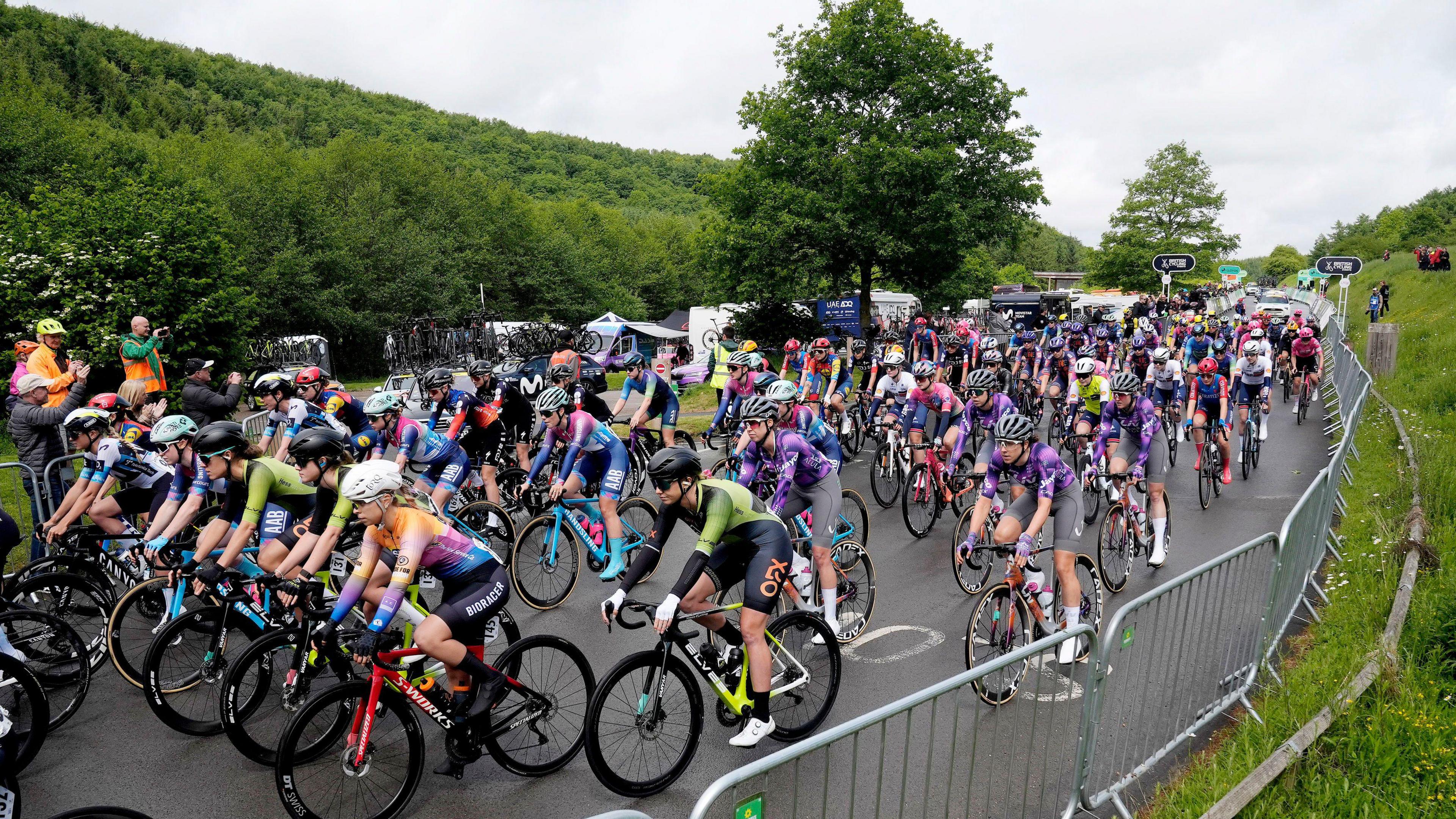 Professional cyclists begin a road race in a rural location. The road s flanked by silver fencing, trees and greenery.