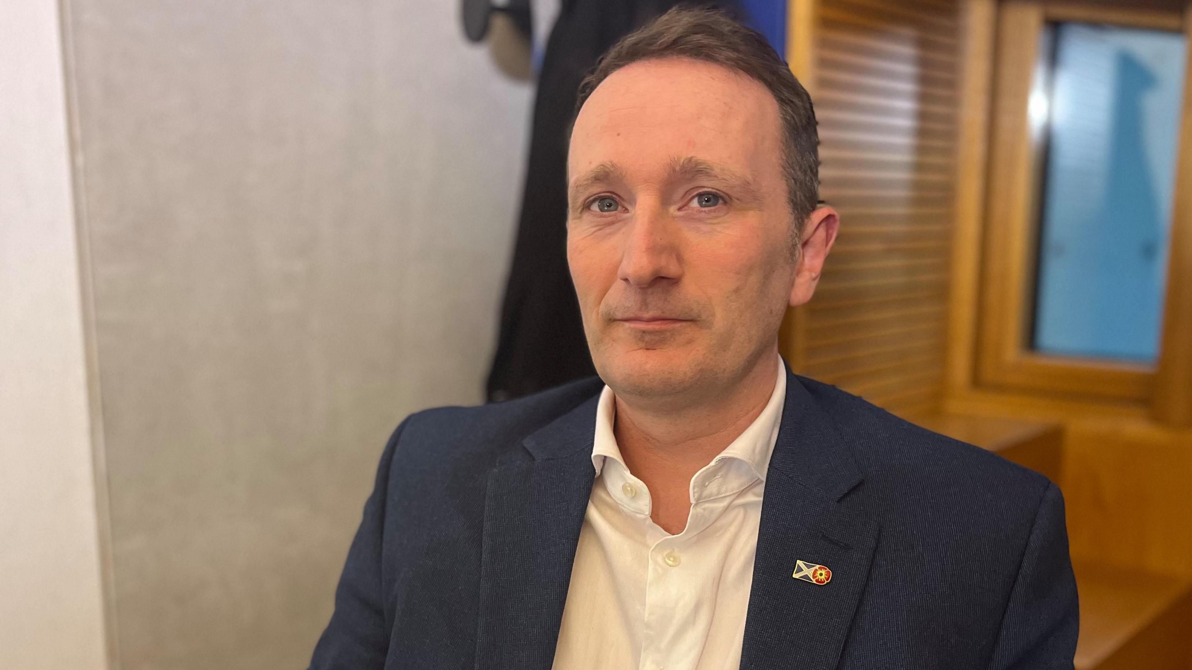 Fulton McGregor sitting in an office at the Scottish Parliament. He is wearing a blue suit and white open-necked shirt, and has short brown hair.