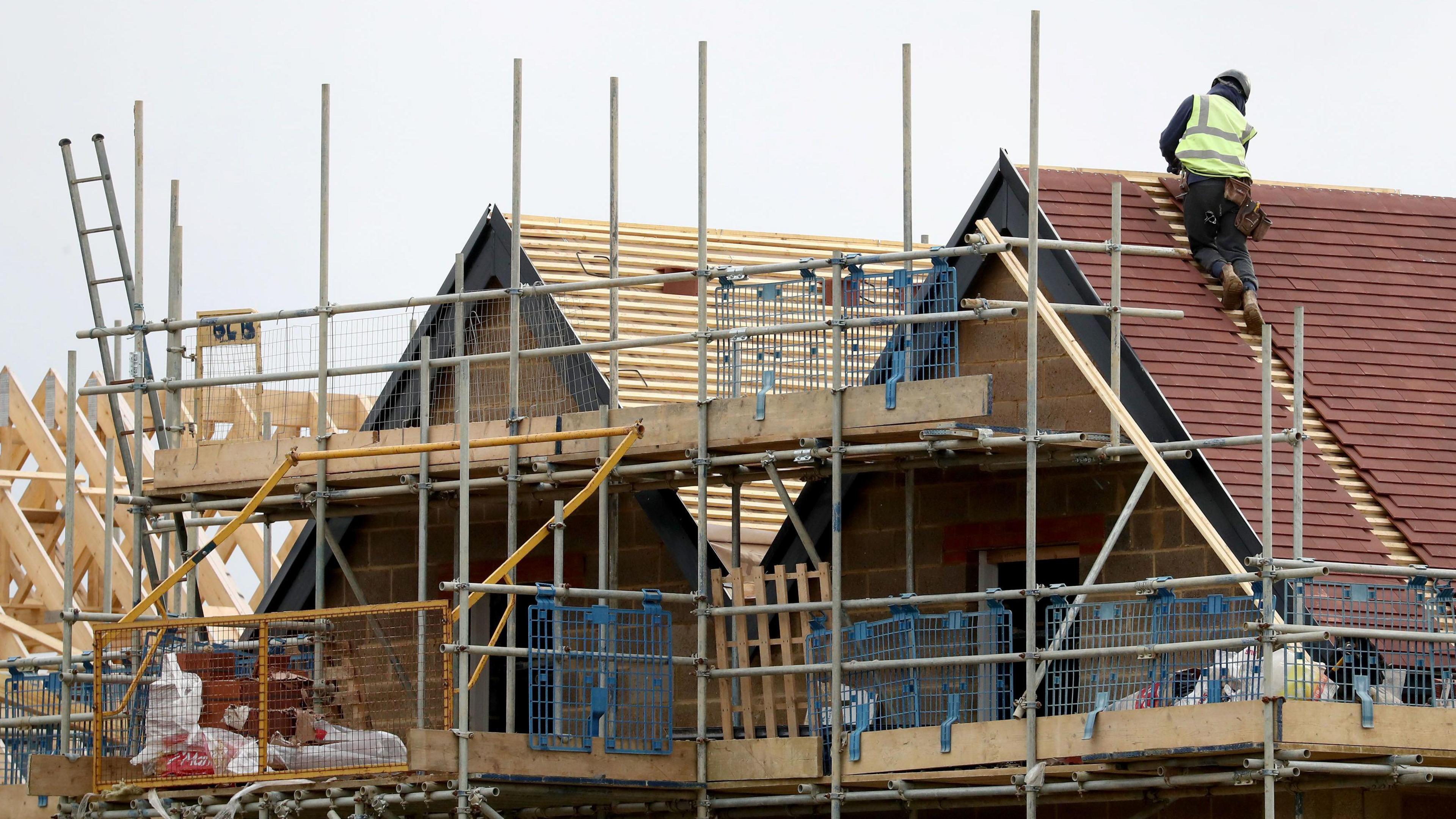 A large domestic property that is partially built, and covered in scaffolding at roof level. On the roof, a worker wearing a high-visibility, fluorescent jacket and hard hat, attends to a task. The weather is clear.