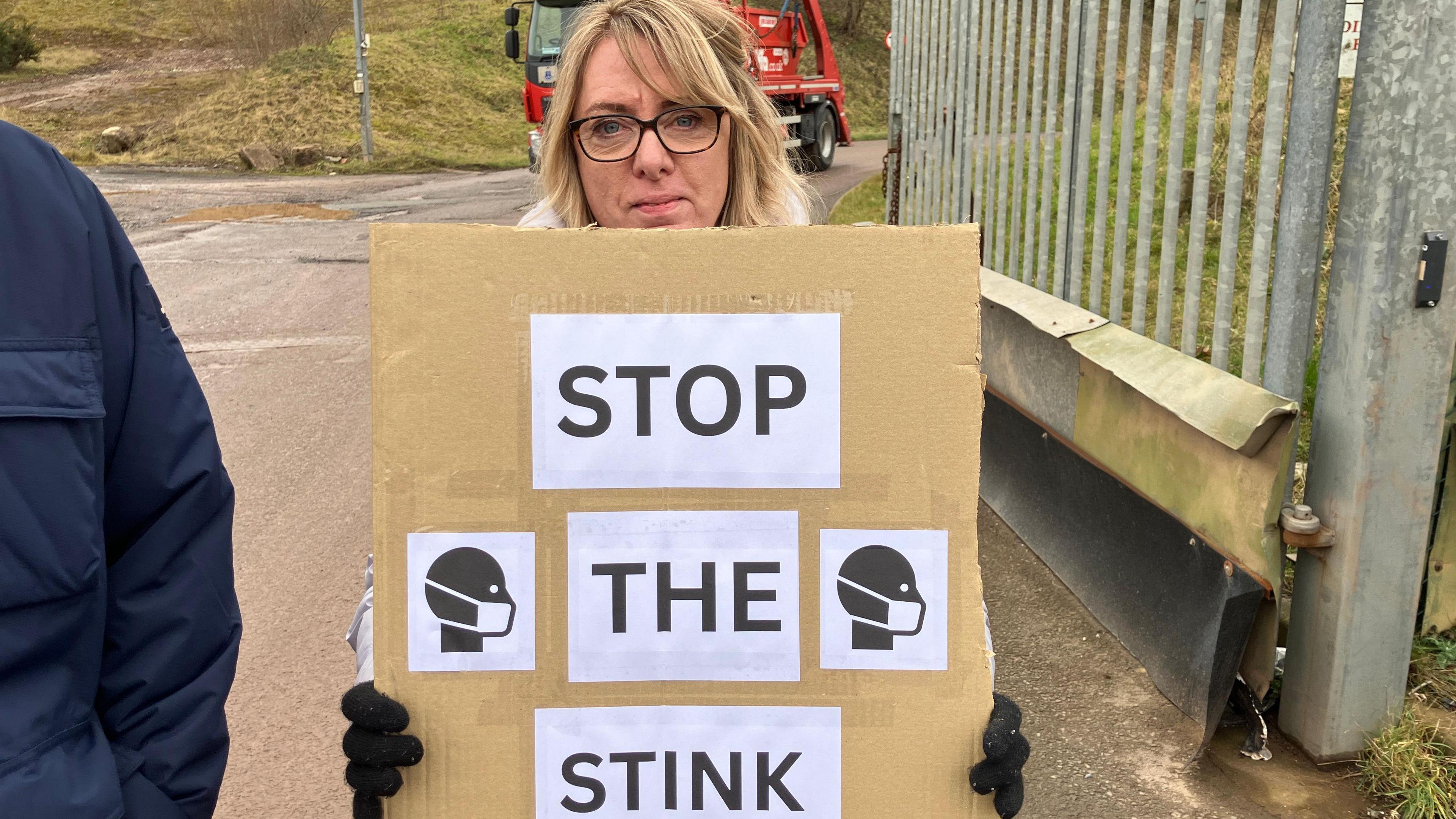 A woman with blonde hair and glasses holds a sign reading "Stop the stink"