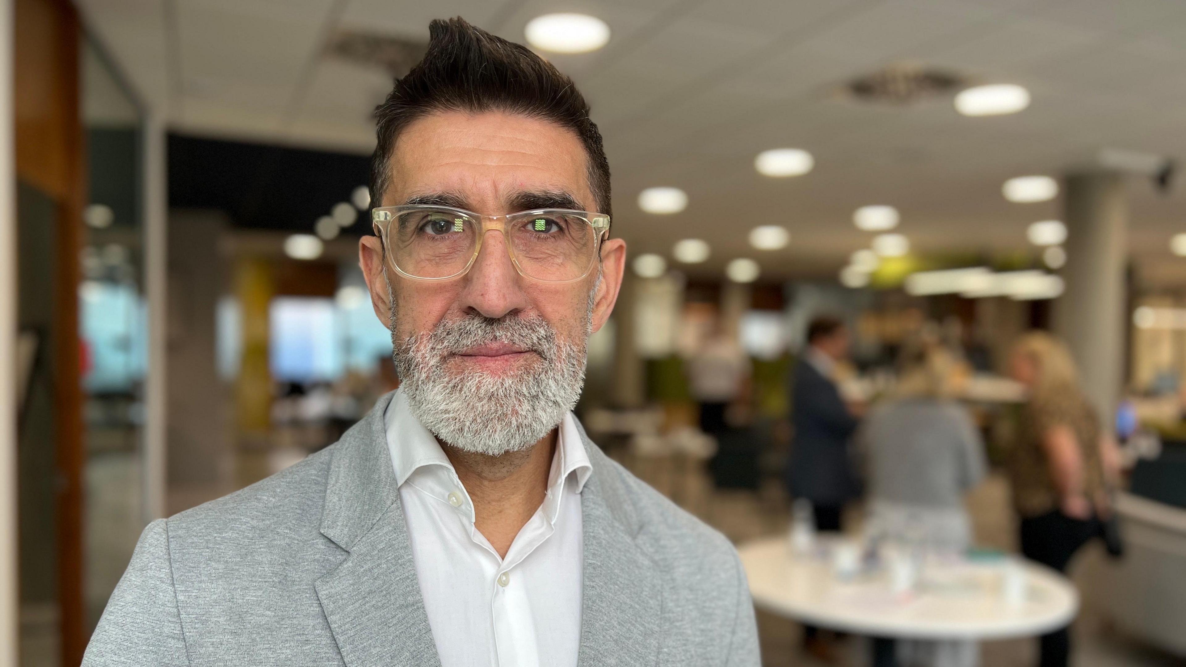 Andrew looks at the camera. He's wearing a grey jacket over a white shirt. He has black hair and brown eyes with a grey, short beard.. He is wearing glasses with a clear frame. In the background - three people are talking to each other by a table.