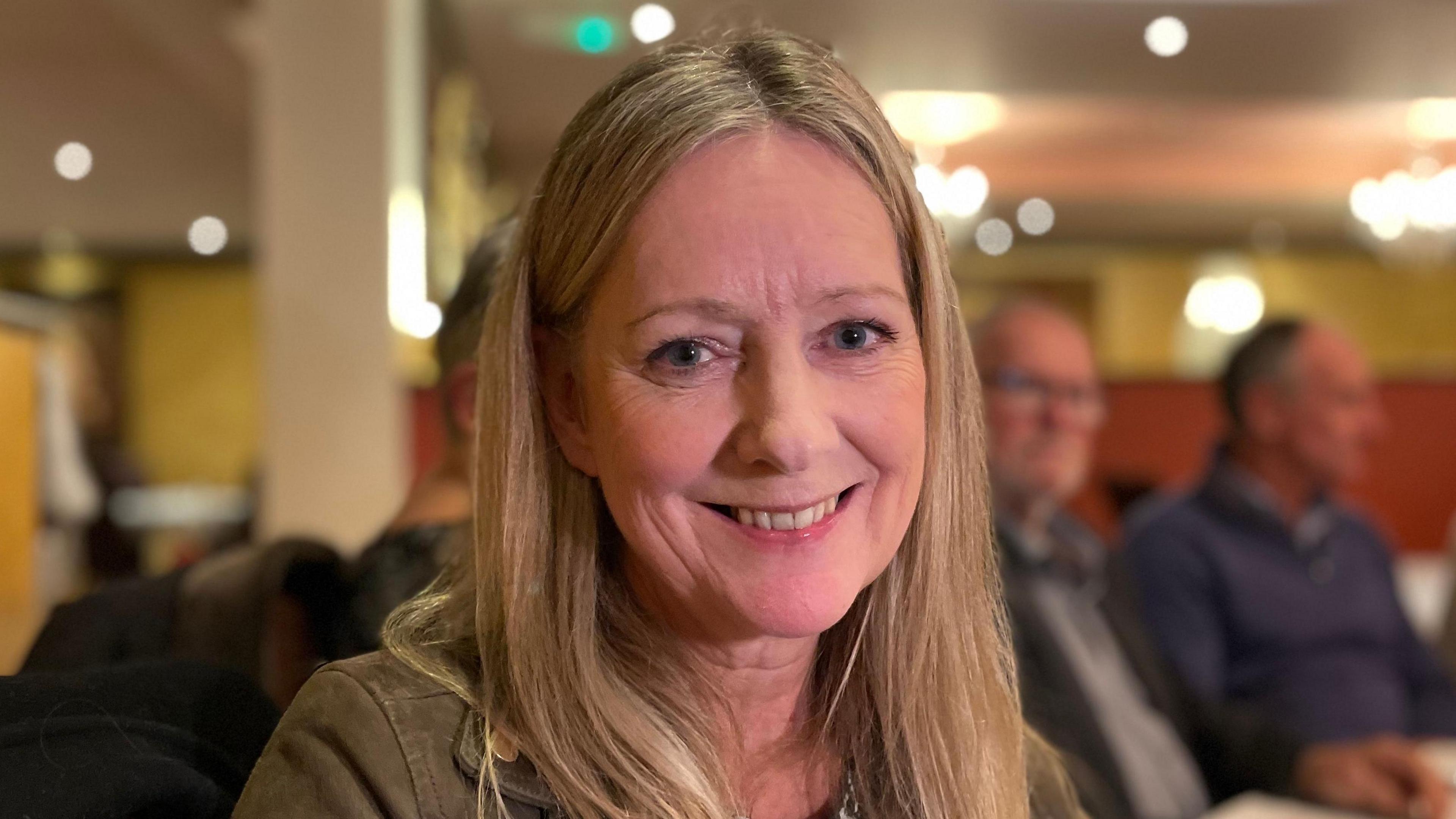 Jacqueline Allen smiling and looking direct to camera. She has blonde, straight hair falling over her shoulders and blue eyes. She is wearing a dark green jacket. Other diners in a restaurant can be seen out of focus in the background. 