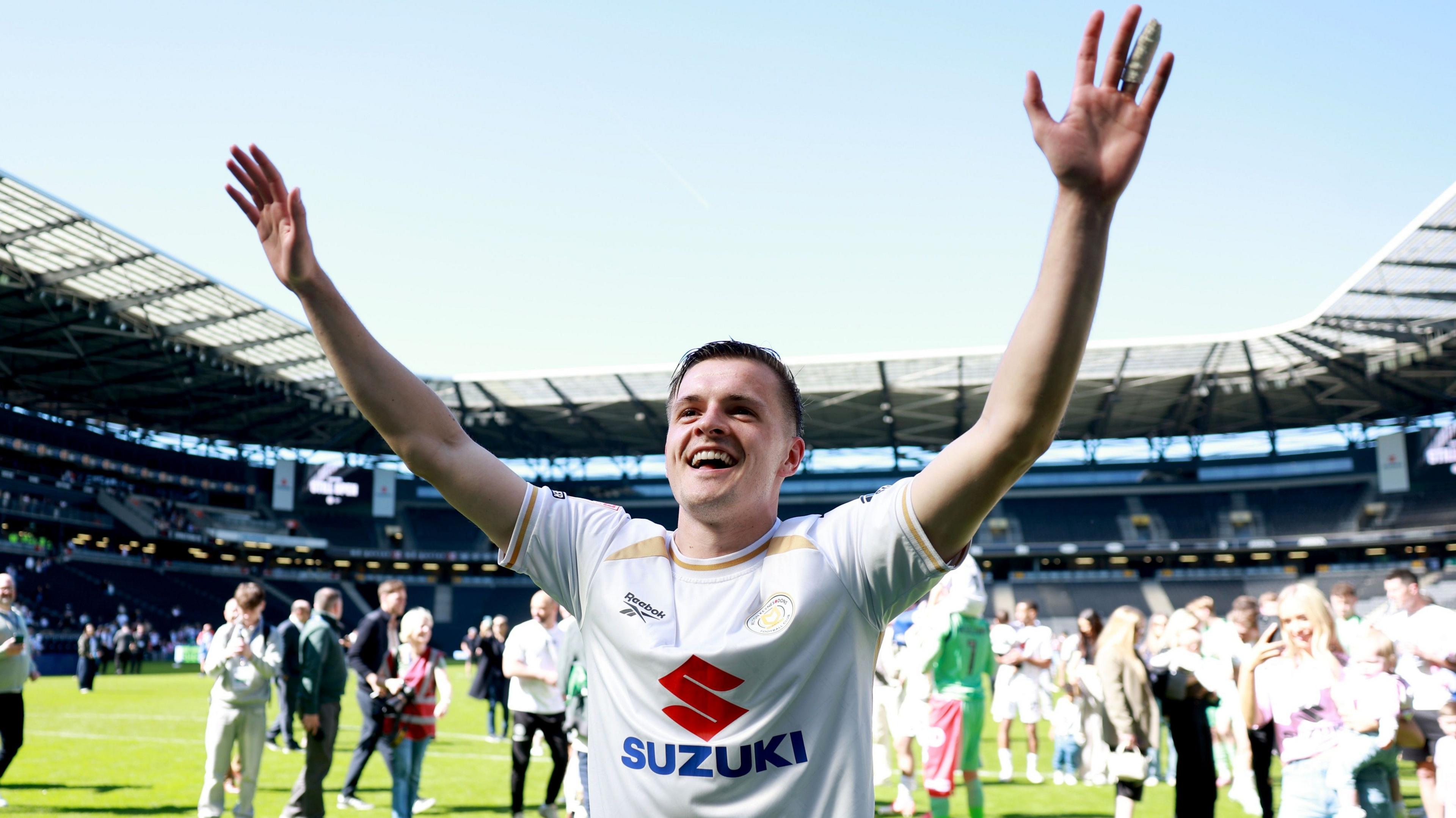 Joe Tomlinson standing looking at a home crowd with both arms aloft and huge smile, wearing MK Dons home kit