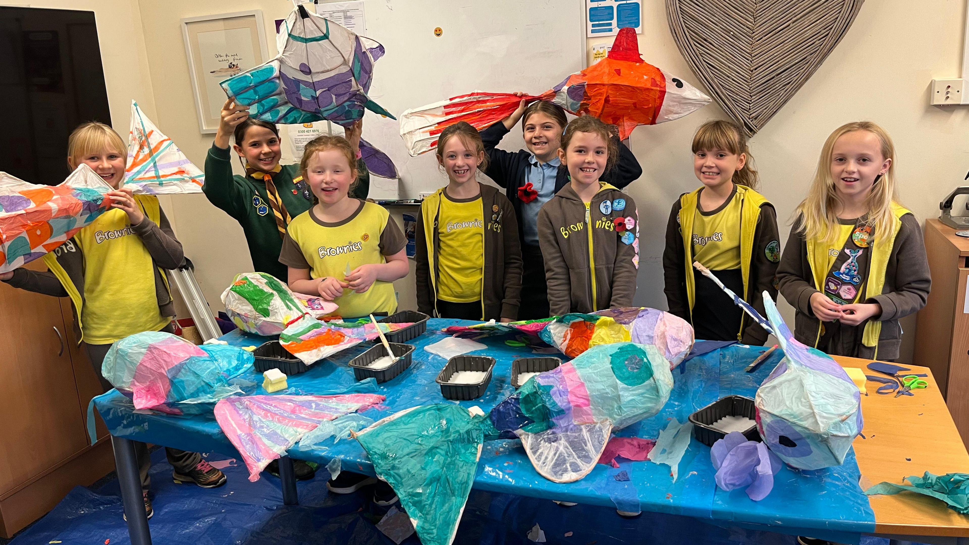 A group of young female pupils stand in a line, wearing brown and yellow brownies uniform. In front of them lies a table covered in colourful fish-shaped lanterns, along with black trays filled with PVA glue and spreaders.