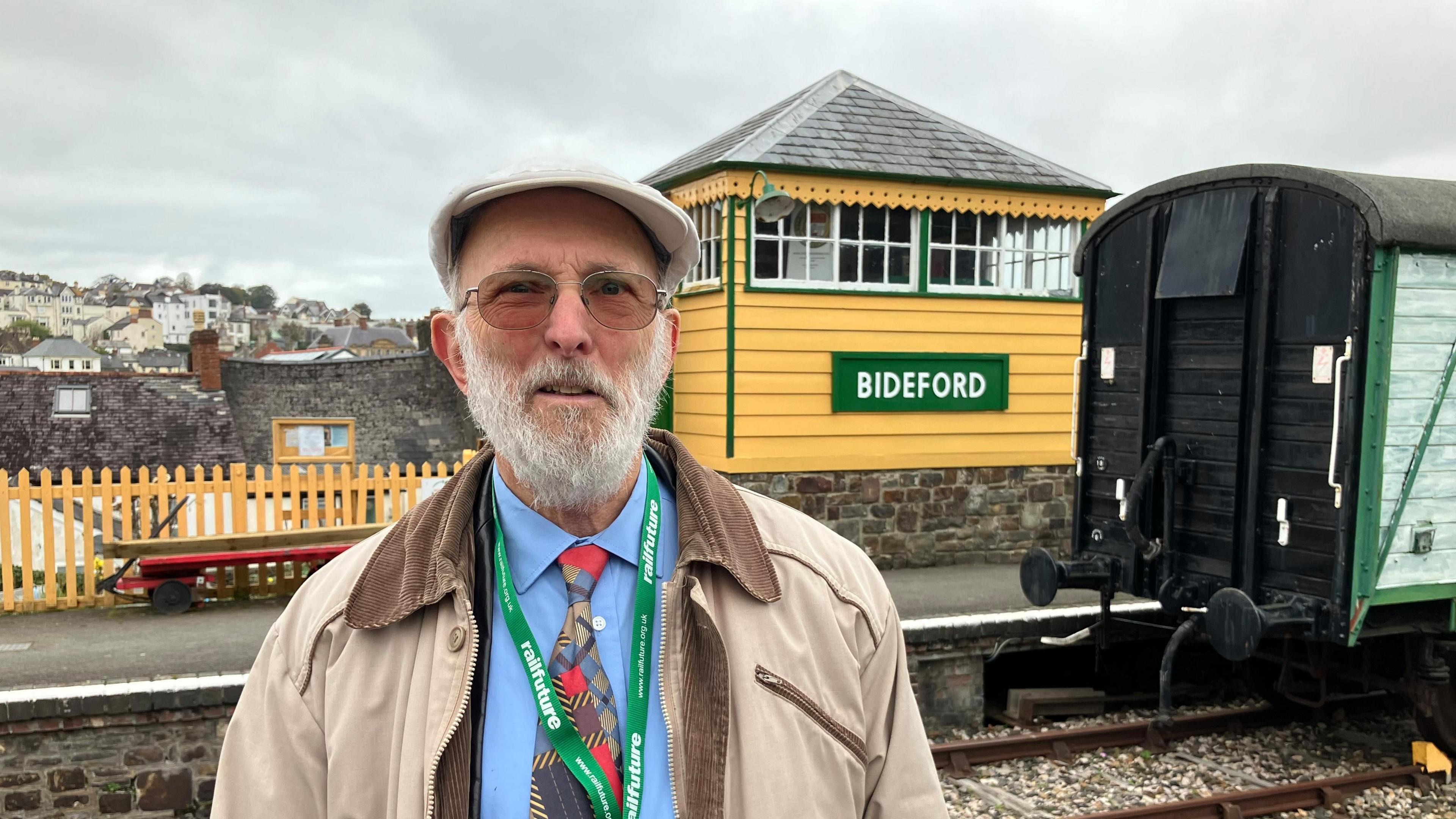 Roger Blake, a grey bearded man wearing a beige jacket, beige cap, blue shirt and sunglasses, stands in front of a yellow railway signal box, which has a sign saying "Bideford" on the front. The back of a railway freight carriage is on the right.