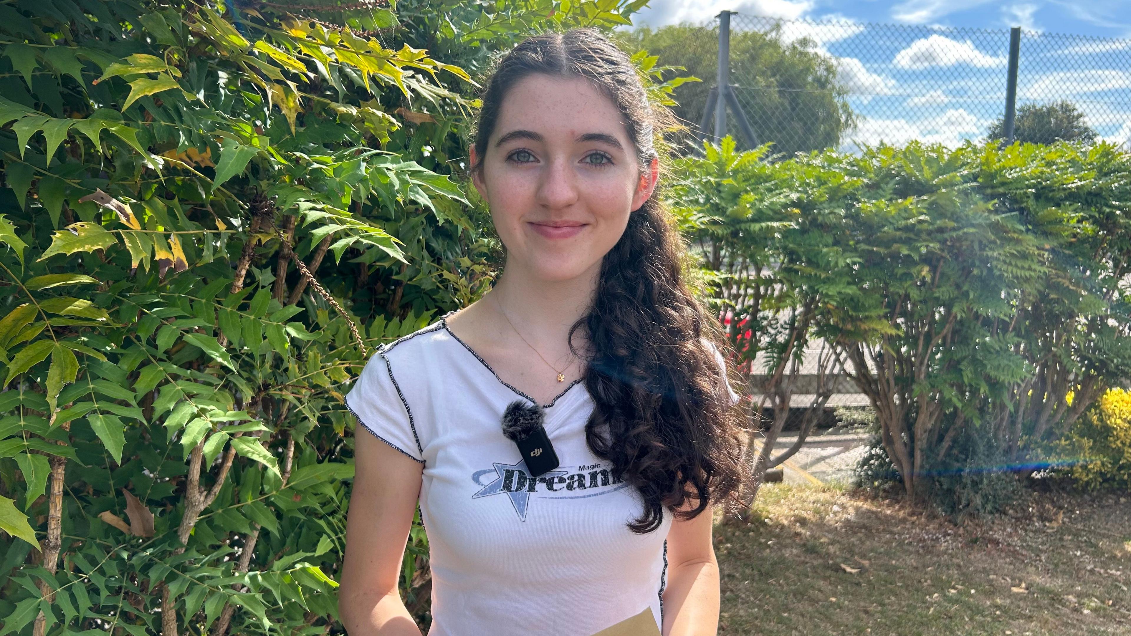 Miranda Fay wearing a white t-shirt which says Dreams across the chest and a mic clipped on the collar. She has long, thick, curly brown hair which is styled in a ponytail resting on her shoulder. She is standing in front of a metal fence and bushes, smiling at the camera.