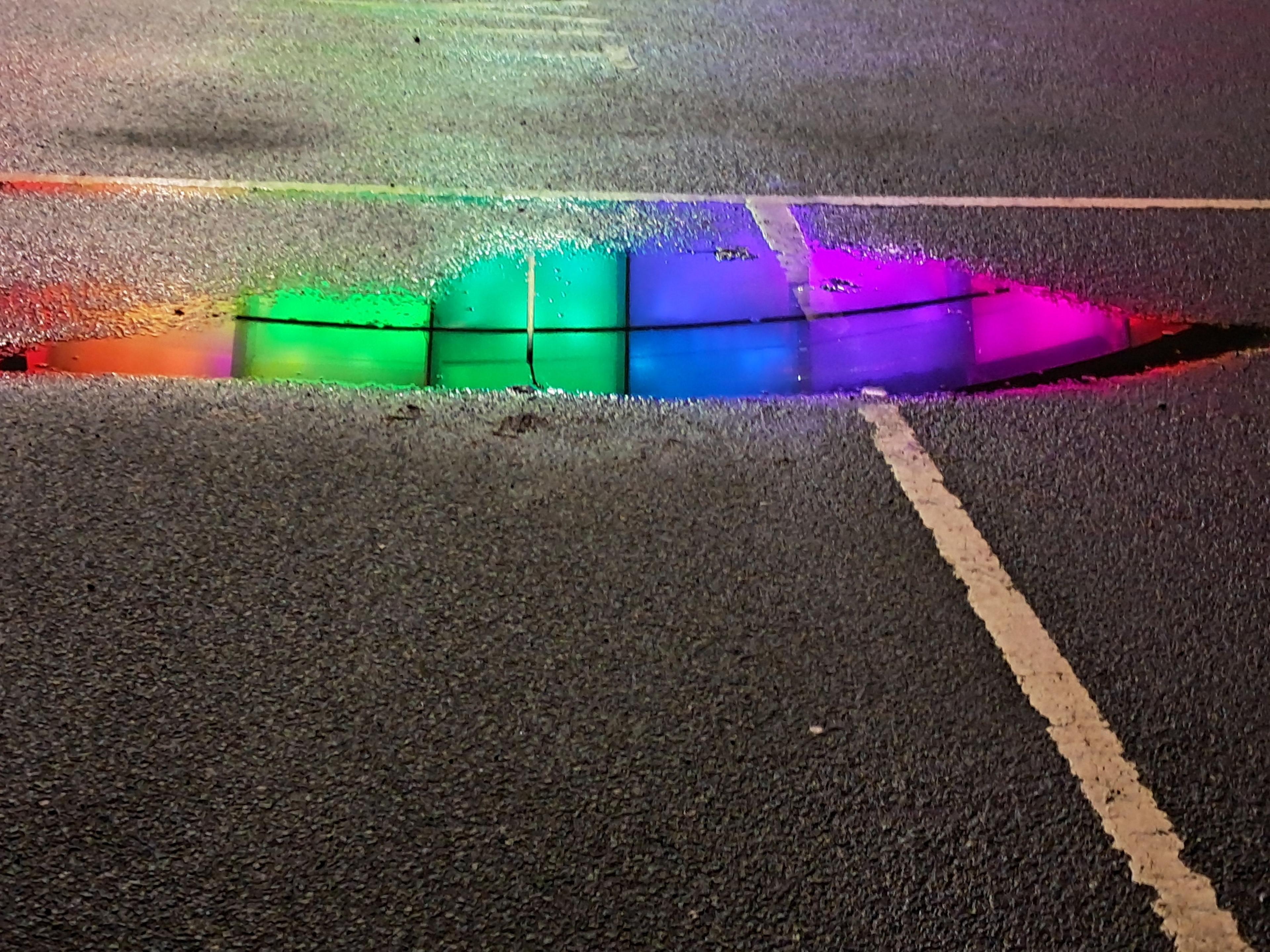 Small puddle of rain water in a car park with the rainbow colours lighting up the Hydro reflected in the puddle