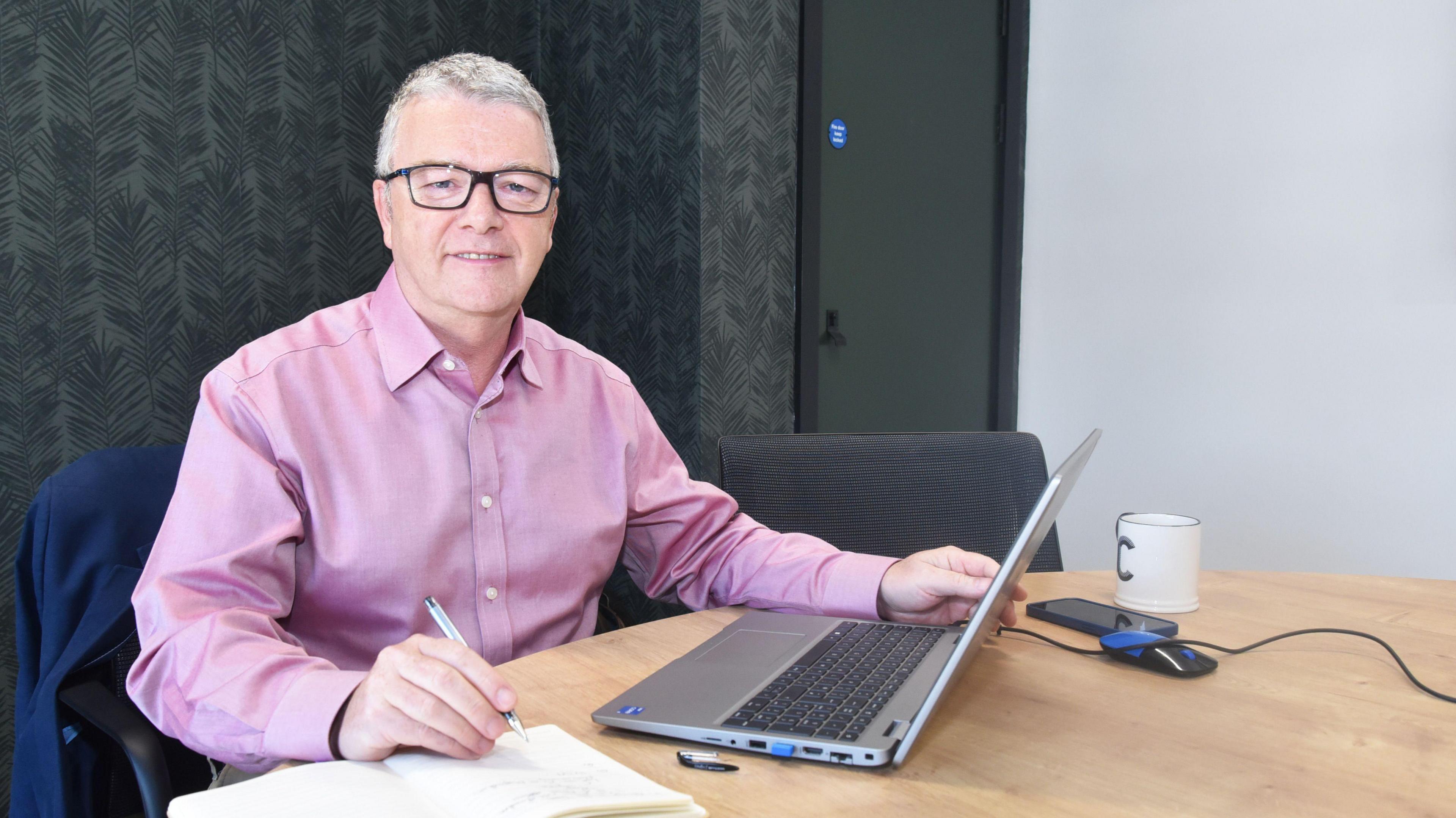 A photo of Clive Heaphy, a grey haired man with glasses and a pink shirt sitting in front of a laptop, smiling at the camera.