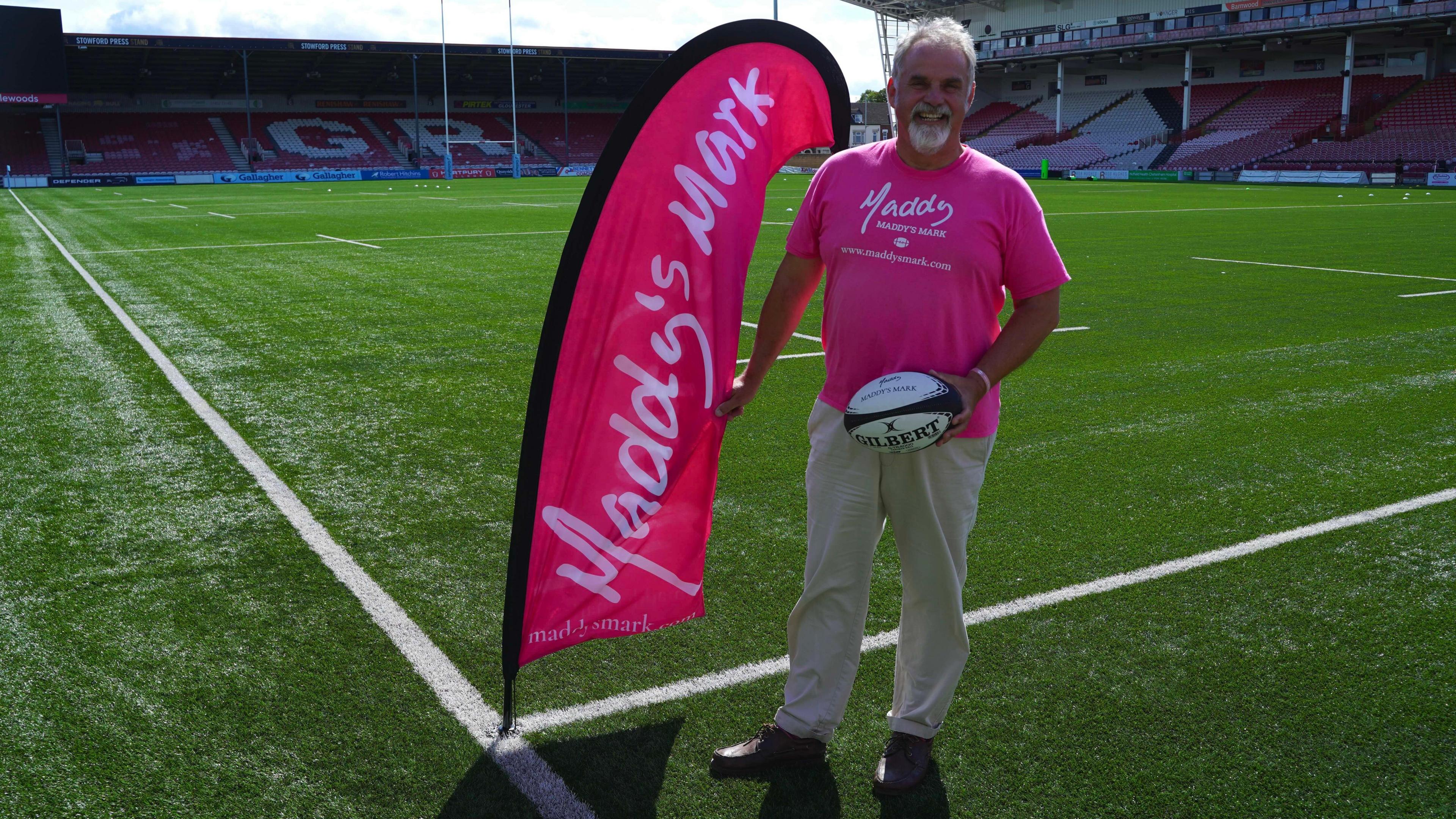 A man standing on a rugby pitch next to a pink flag with Maddy's Mark written on it in large white letters. He has short white hair and facial hair and is wearing a bright pink shirt with the same logo on it. He is holding a rugby ball. He is looking at the camera and smiling.