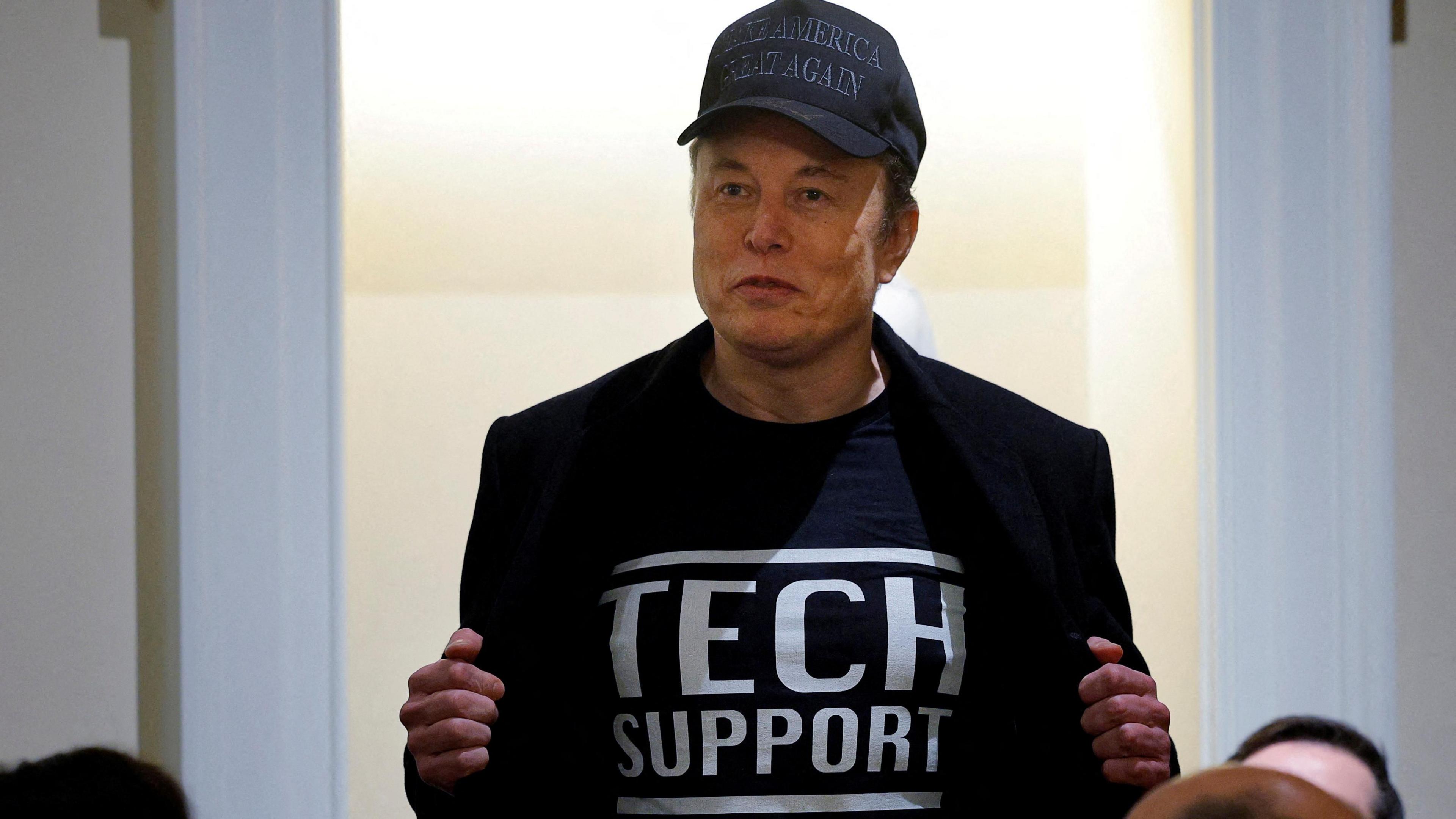 Elon Musk, wearing a blue baseball cap reading Make America Great Again, holds his jacket open to reveal the Tech Support slogan on his T-shirt.