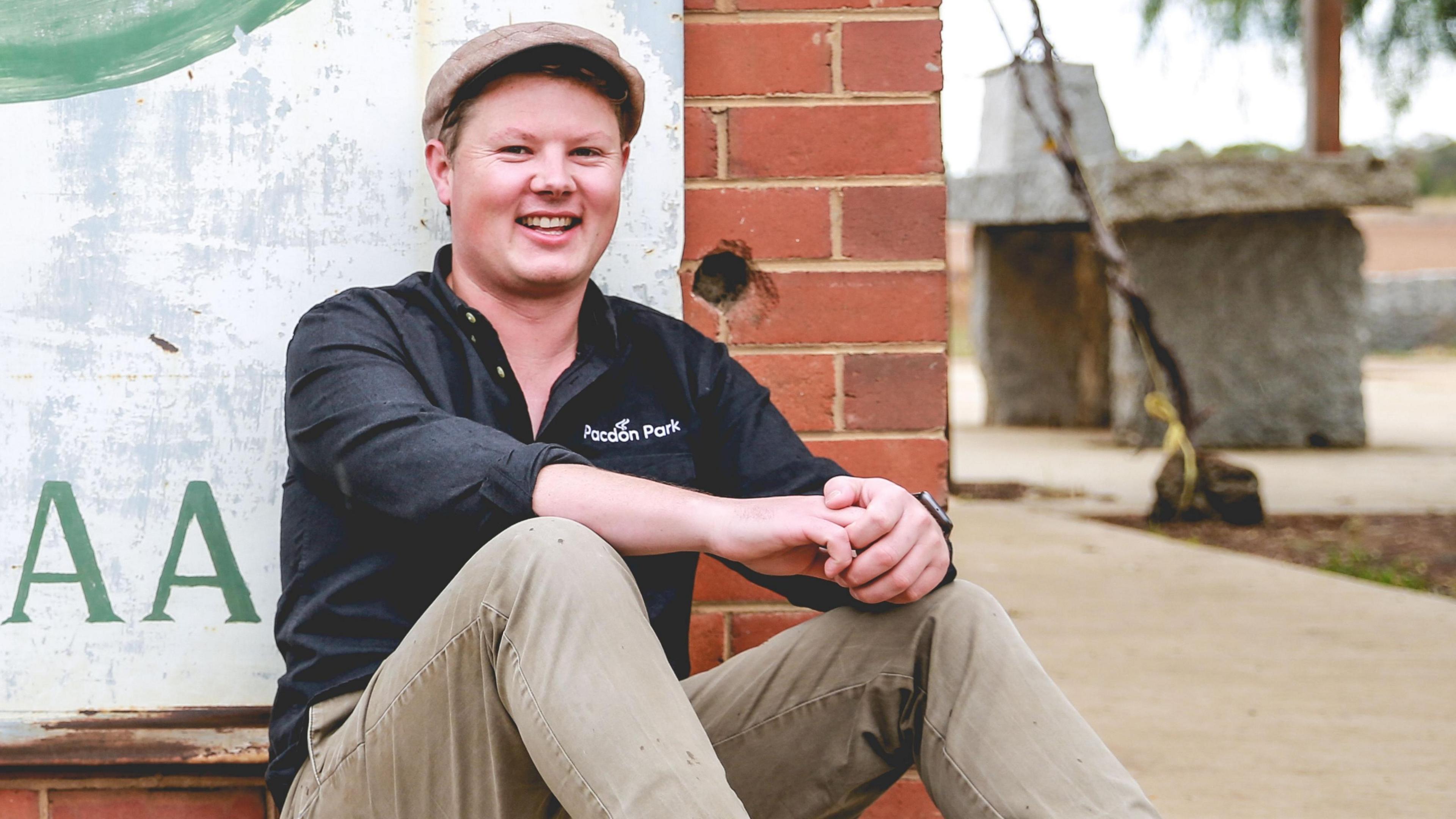 40-year-old Jim Arrowsmith smiles sitting on the floor against a brick wall. He is wearing a cap and a black shirt with the words "Pacdon Park" written on the front.