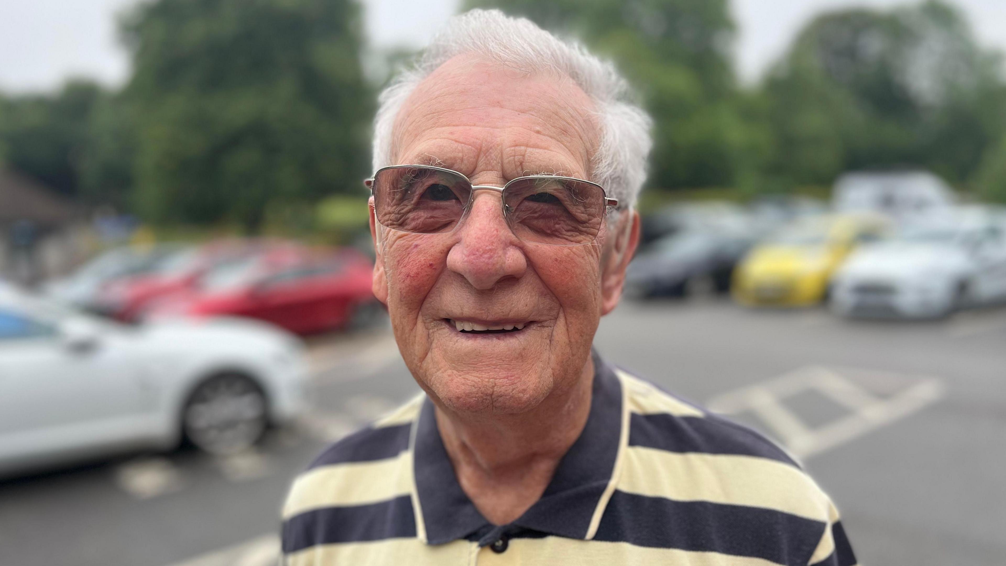 Alvin Trevenna is wearing sunglasses and facing the camera while wearing a lemon and navy striped polo short, with blurred out cars in a car park visible behind him.