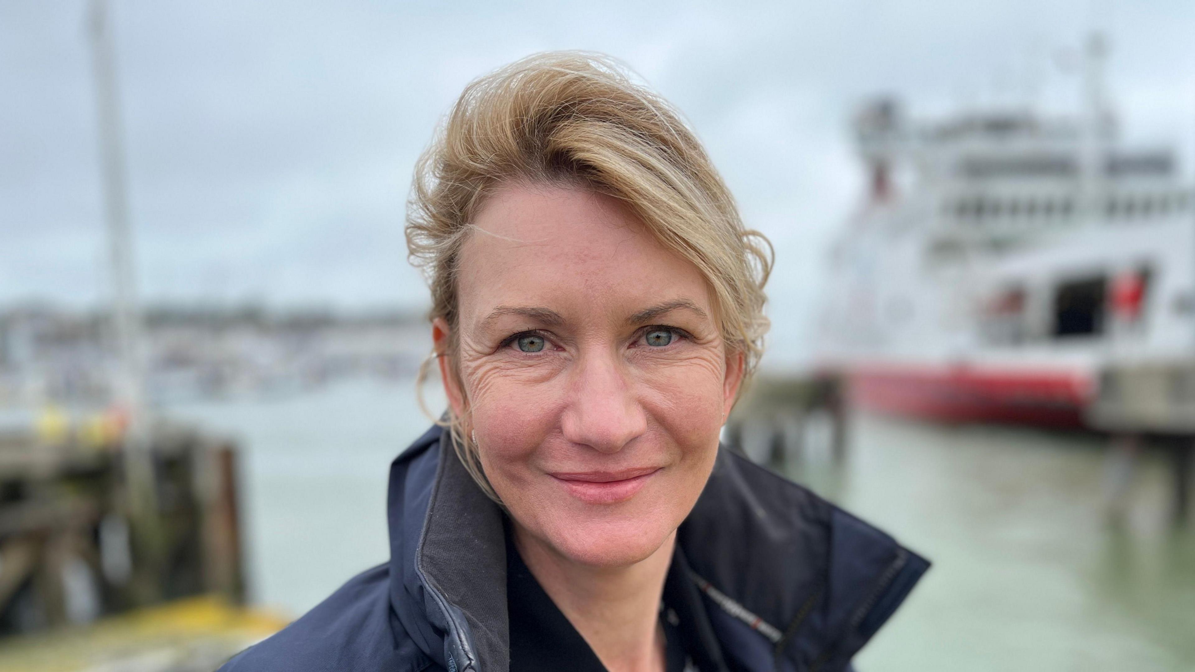 A blonde haired woman from the shoulders up, with a harbour out of focus in the background