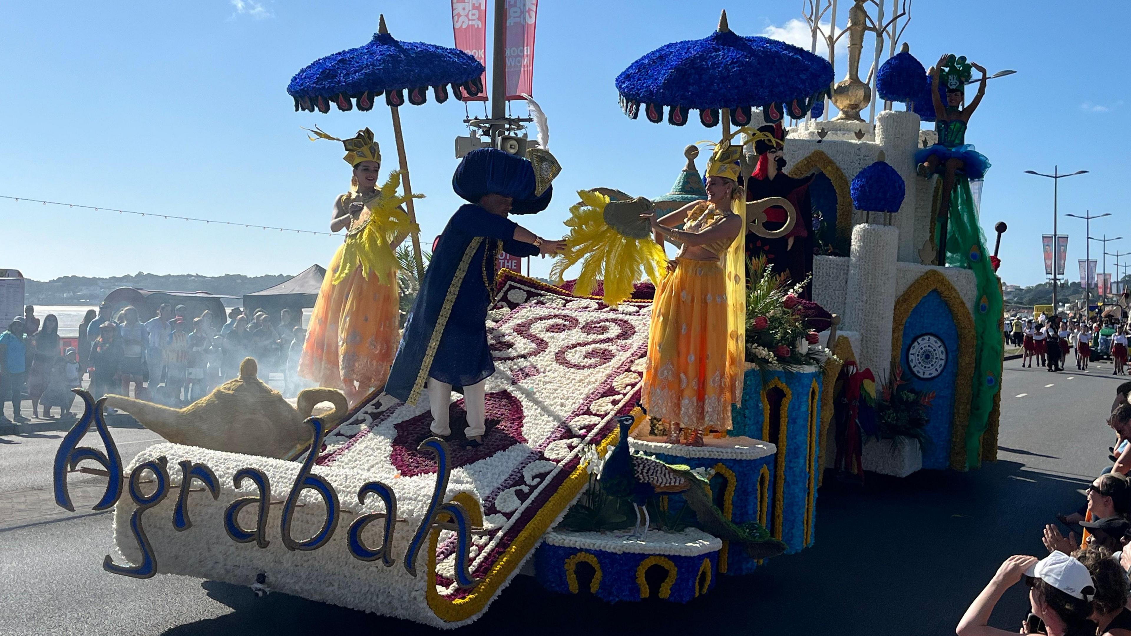 A float in the Battle of Flowers parade which holds two women dressed in golden gowns wearing crowns and holding parasols and a man dressed in blue wearing an extravagant hat. The float features a Middle Eastern style castle with blue domes atop towers towards the back of it    