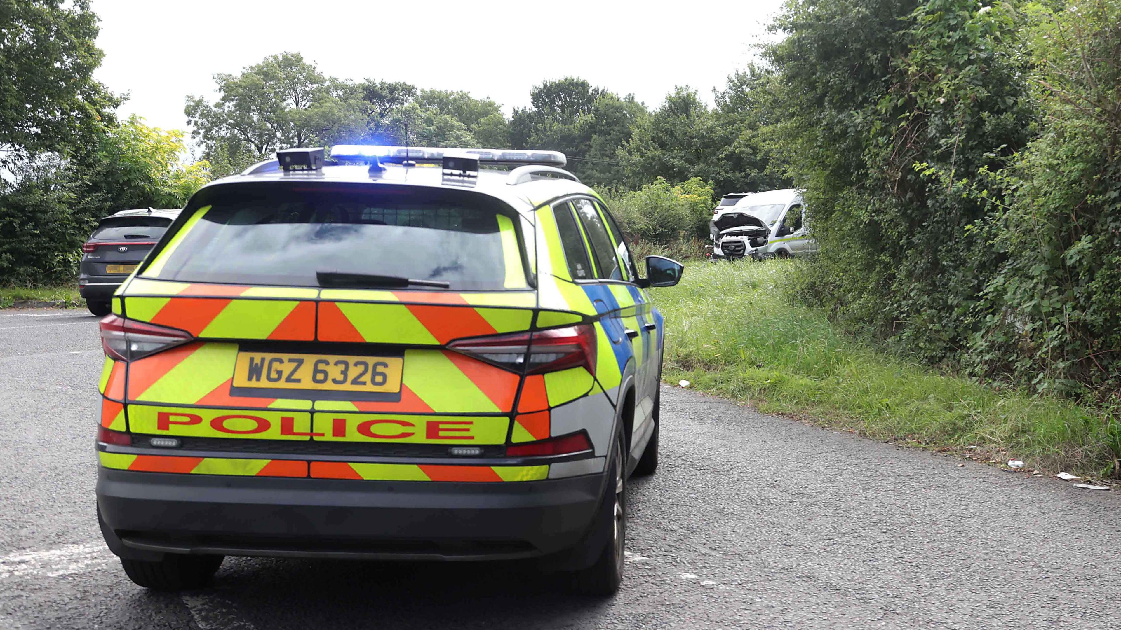 A police car with a car in the distance and a white van also in the distance. There is grass and trees along the side of the road.