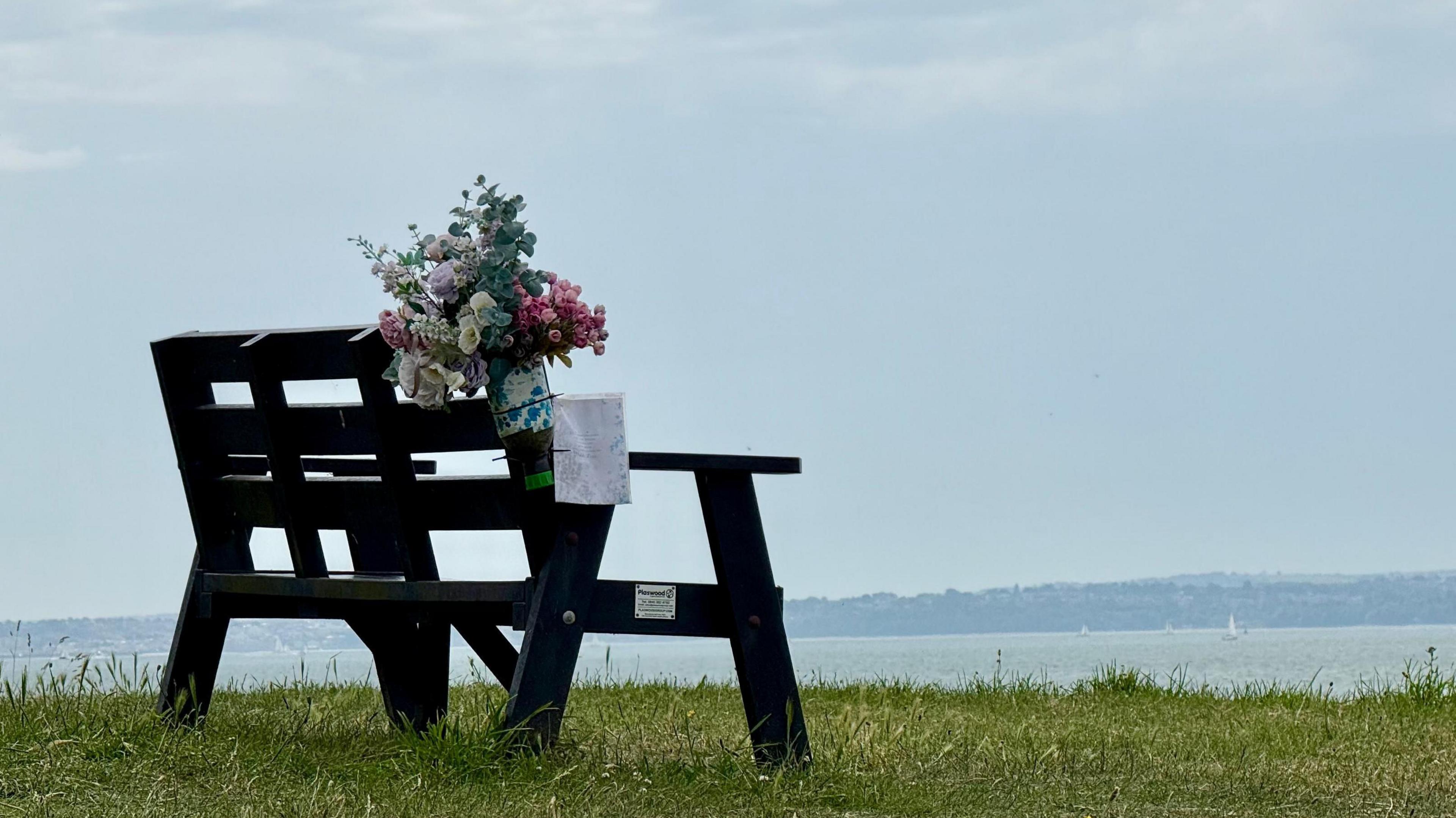 An empty bench sits on grass with a view over water towards land in the far distance. A small yacht can be seen in full sail. A bunch of flowers with a note is attached to the back of the bench.