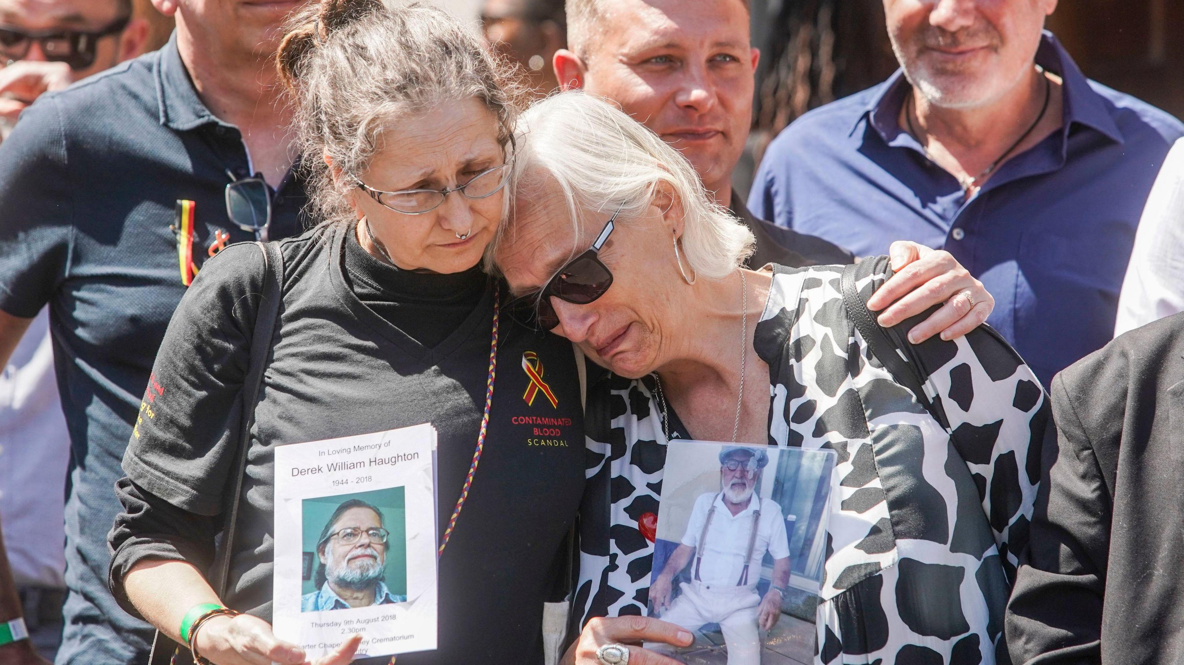 Two women outside the infected blood inquiry comfort each other. One, with white hair and sunglasses and a black and white blouse, leans on the shoulder of the other, who has brown hair and glasses and a black top. They are each holding photographs of a man with a white/grey beard and glasses.