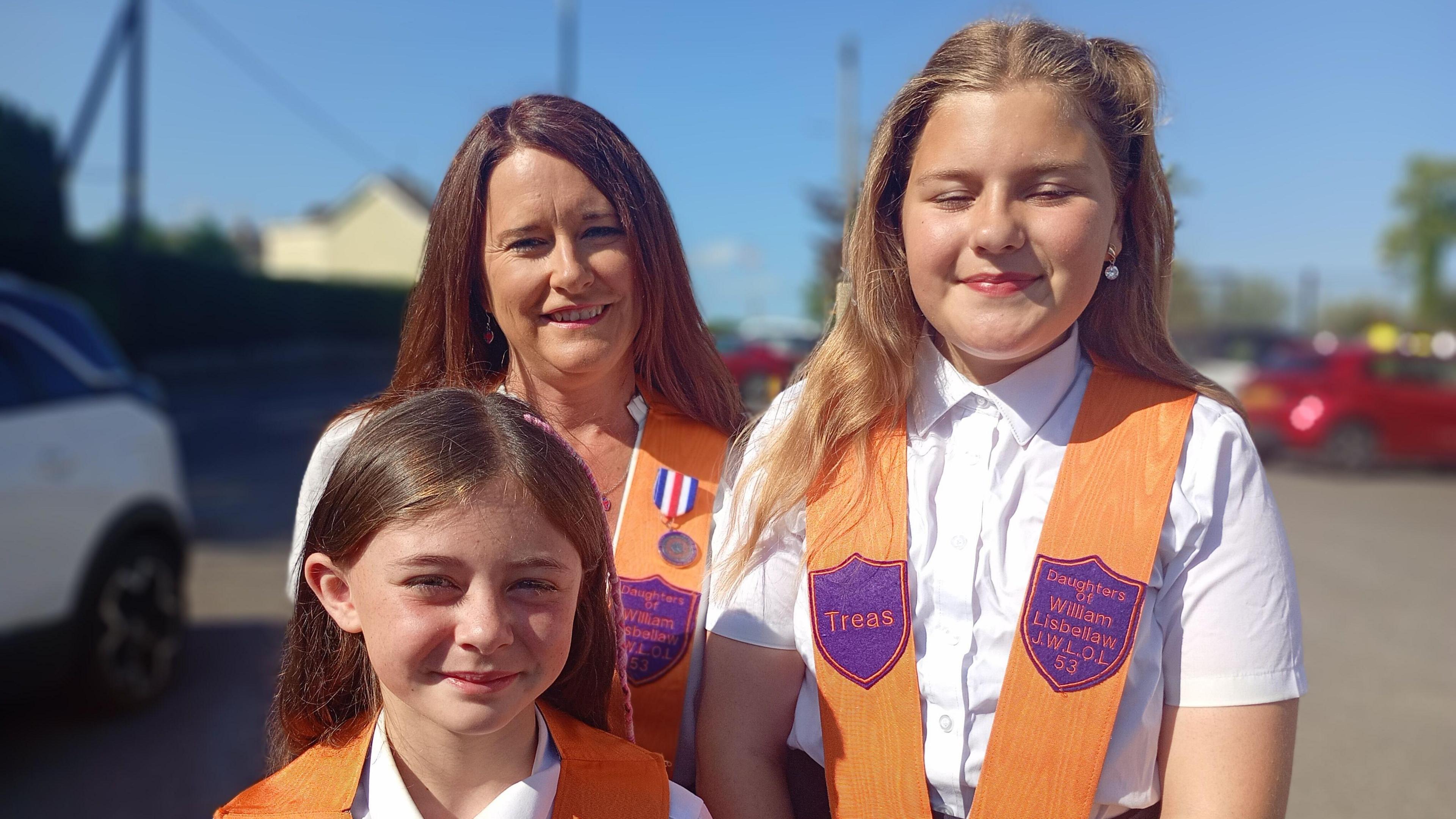 A woman is standing with two girls. All three are wearing bright orange sashes with purple badges. The woman has long dark brown hair while Jessica has shoulder length light brown hair and a white blouse. Alia has brown hair brushed back over her shoulders.