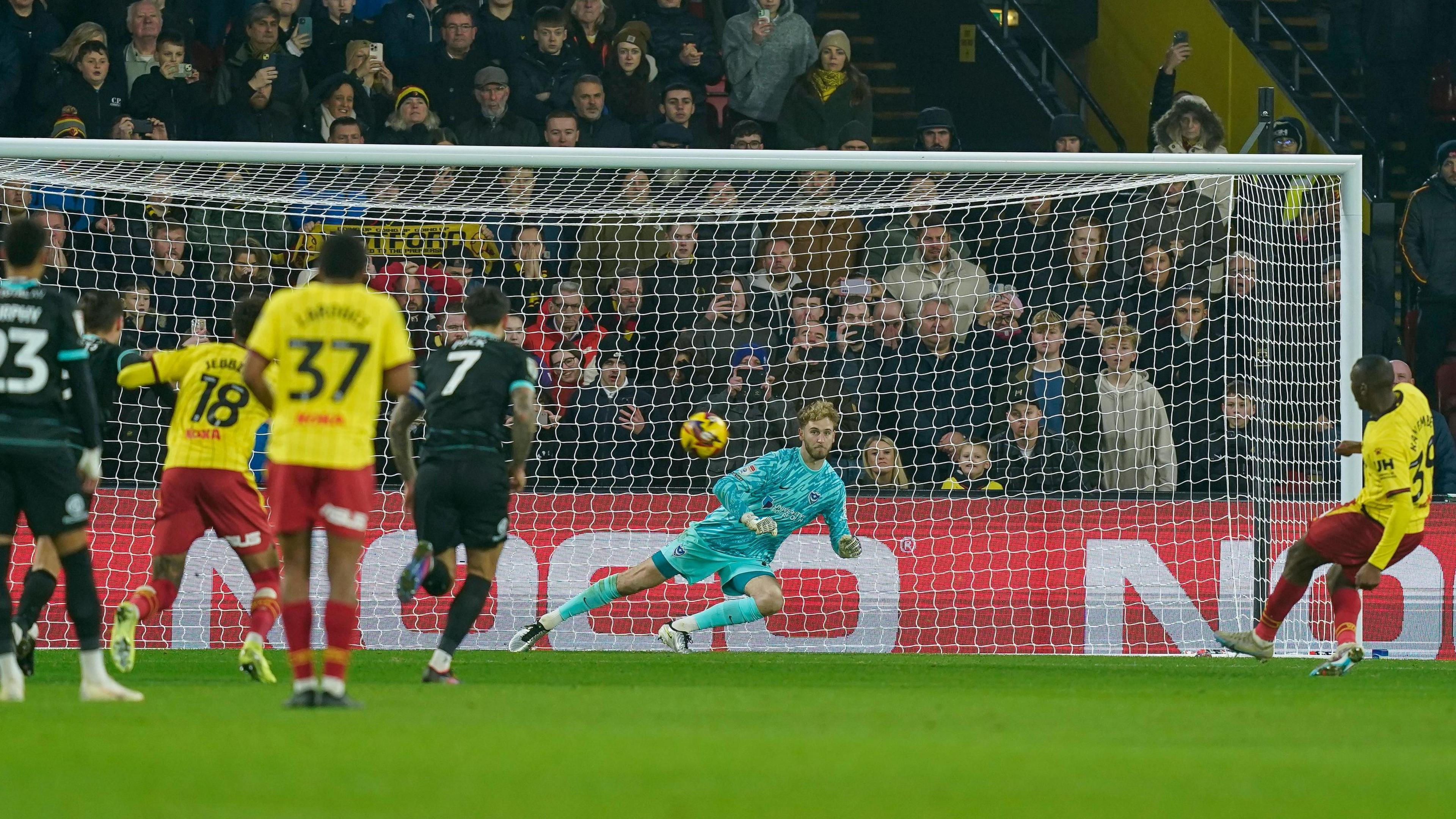 Watford's Edo Kayembe places a left-footed penalty high past Portsmouth's Nicolas Schmid, who dives the other way, into the net to equalise.