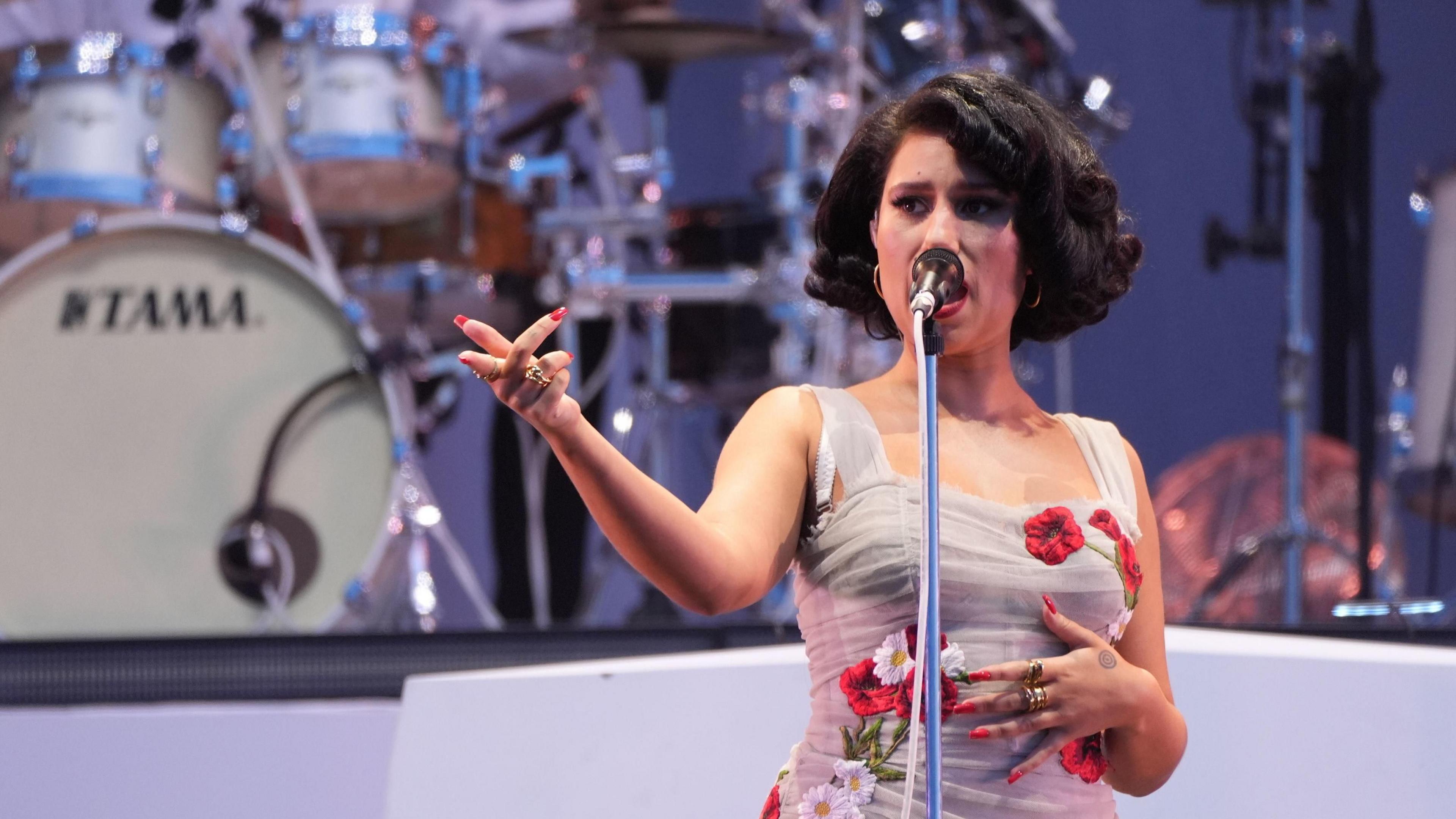 Raye in a flowery dress singing from a microphone and band behind her