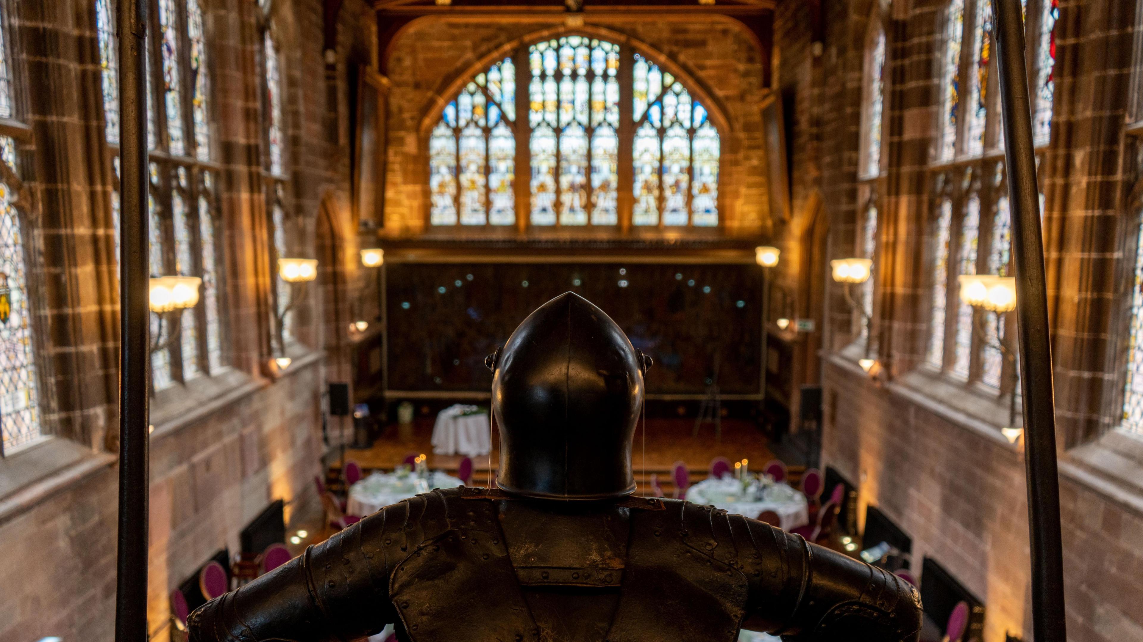 The inside of a medieval guildhall, with high ceilings and stained glass windows on all sides. The picture is taken from up high behind the statue of a knight which you can see in the image looking out into the hall. 