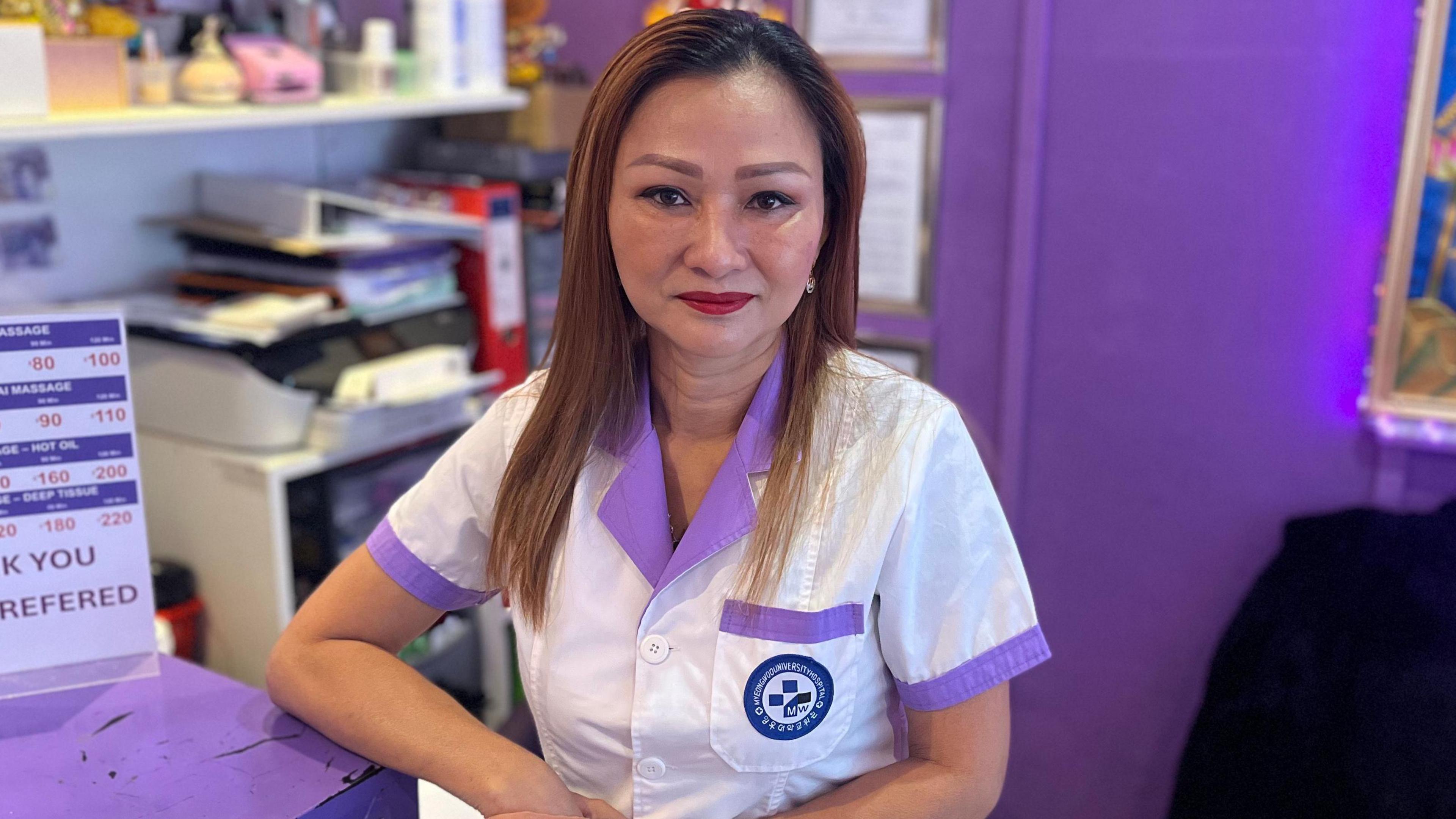 Waraporn Simpson in her work uniform which is a white tunic with purple trim. She is leaning against her reception counter and a purple wall is behind her.