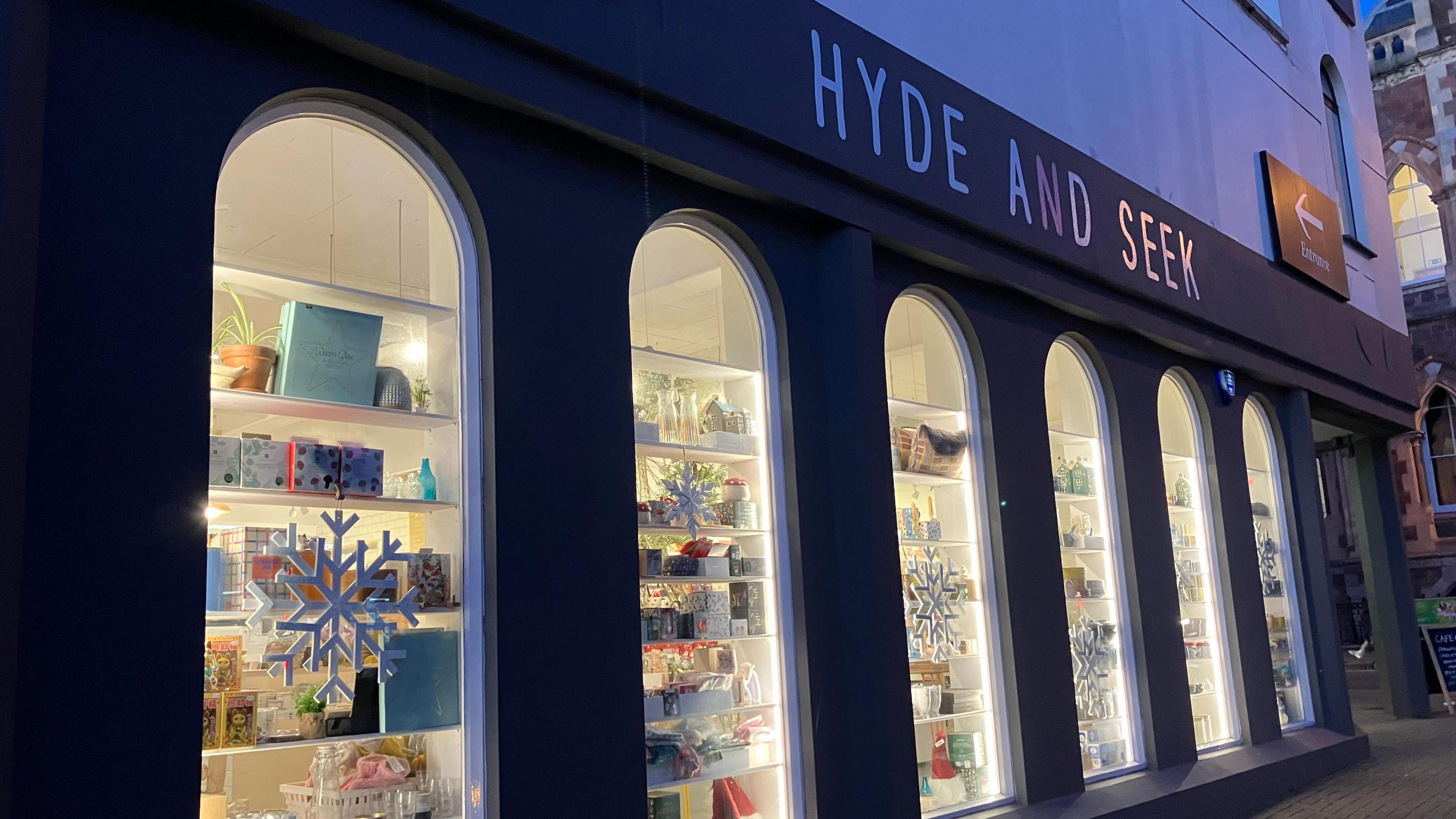 Under the signage of Hyde and Seek, six shop windows can be seen, lit-up as night falls, with stock in each window and snowflakes adorning the glass