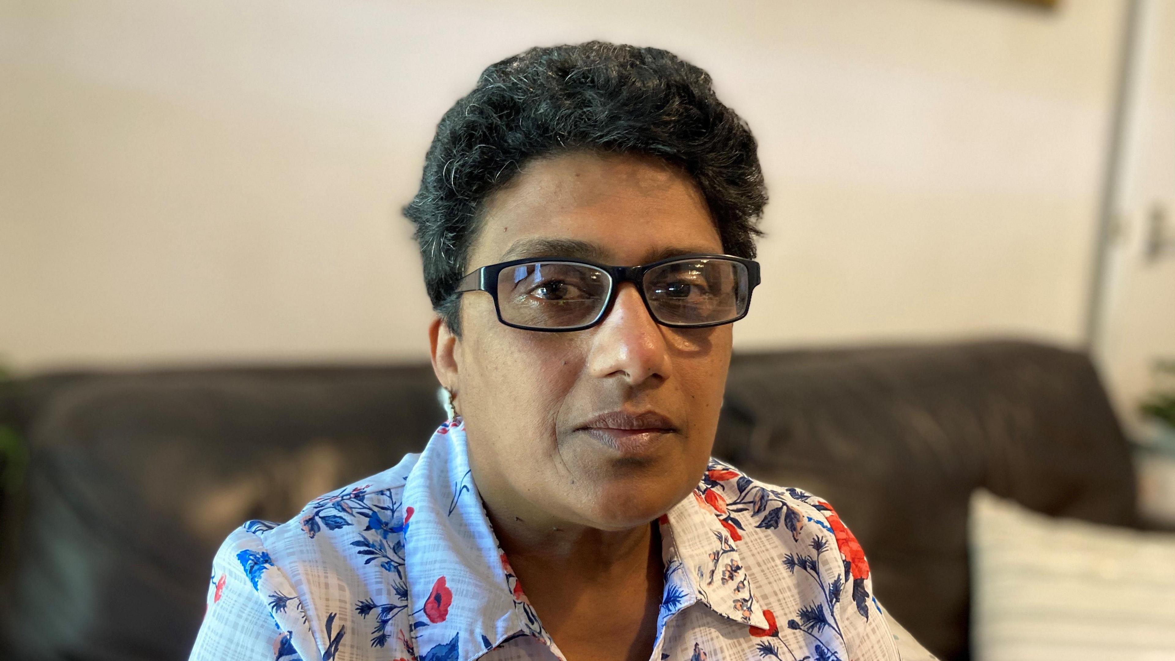 Kumudini Nicola sitting on a sofa in her home. She is wearing glasses and tears are in her eyes.