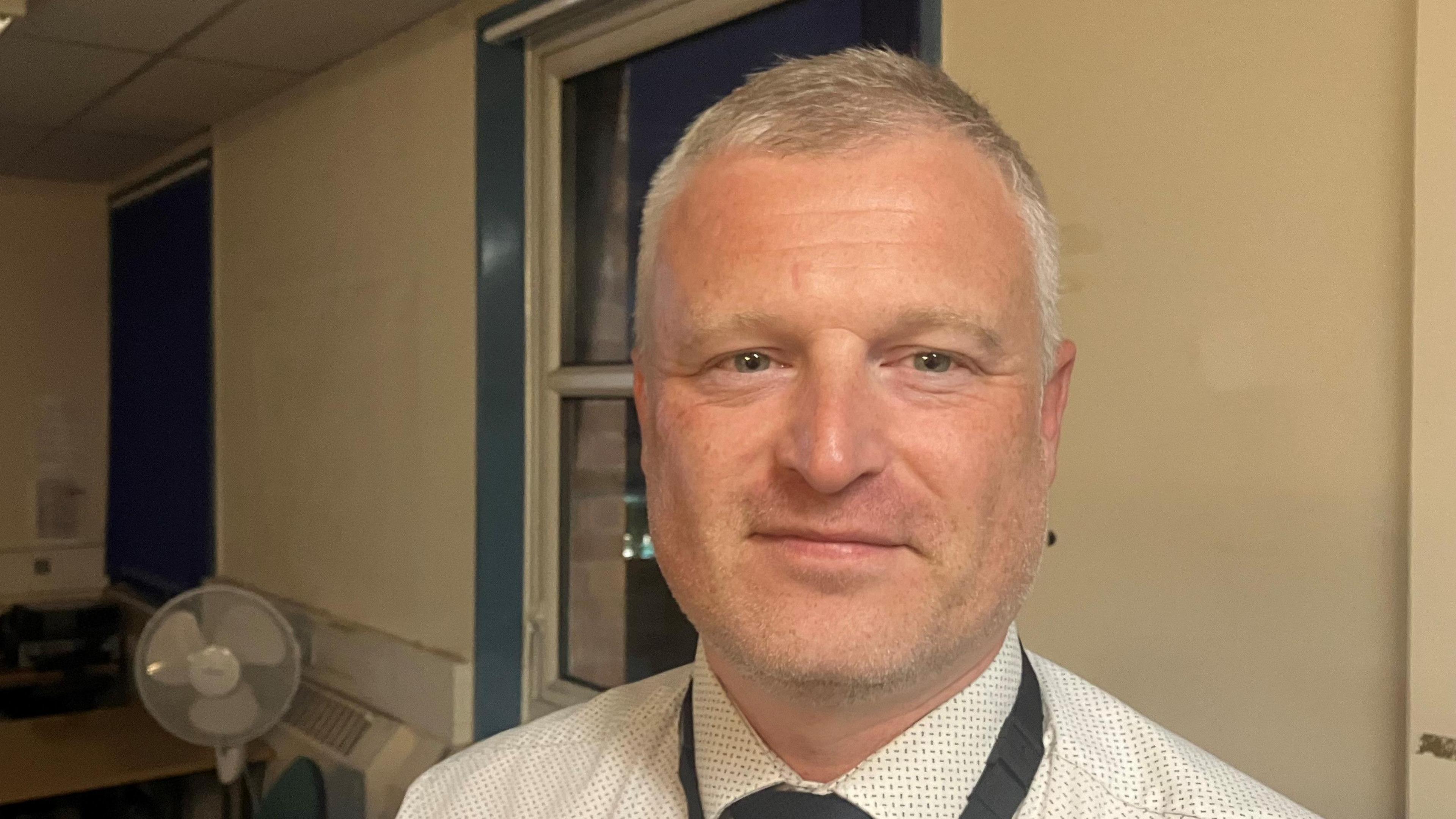 Senior police officer at Greenheys Police Station in Moss Side in a police office room wearing  white shirt and a black tie, with a black lanyard. 