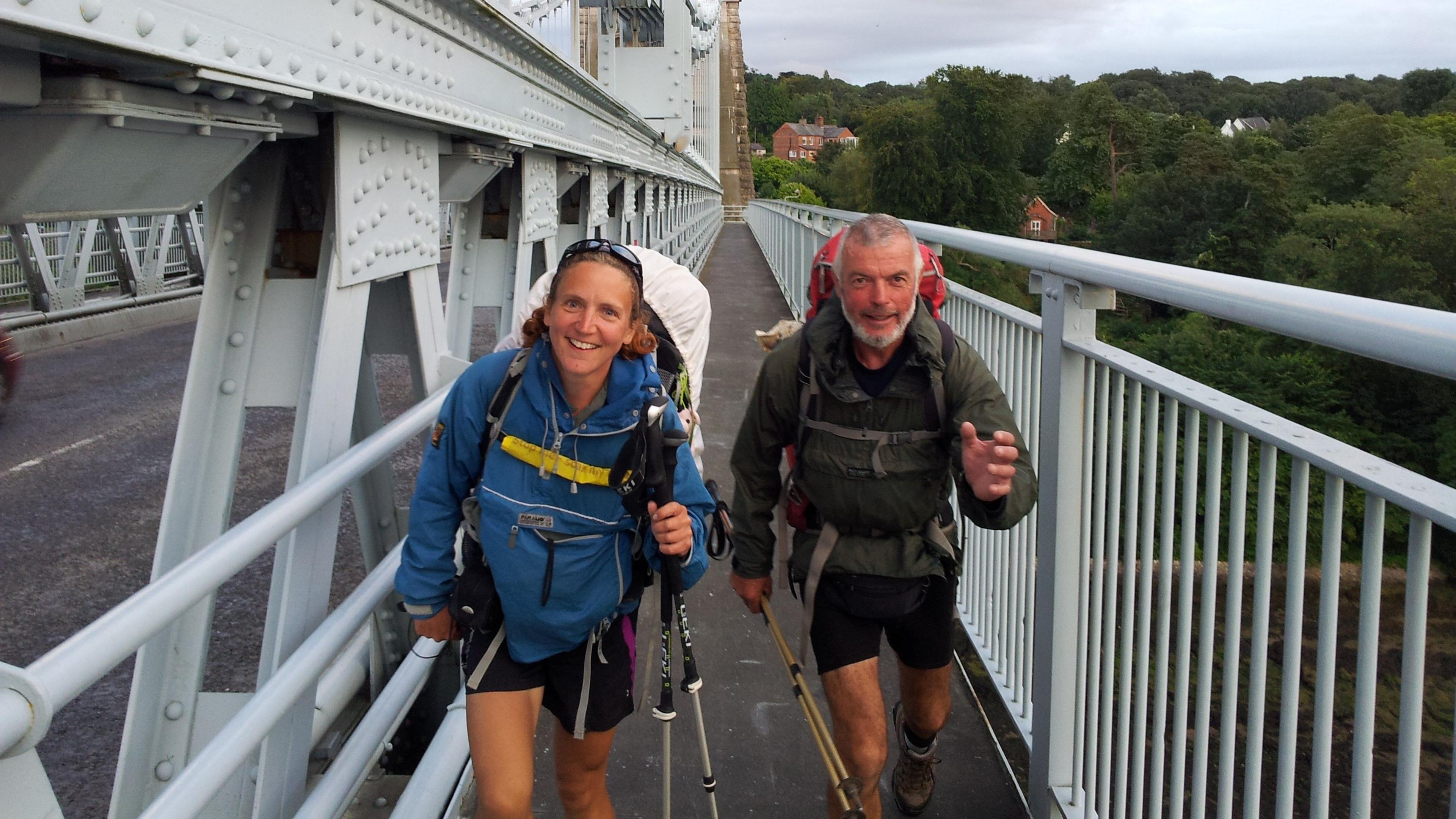 Zoe, wearing a blue waterproof coat and shorts, and Mike, in a green waterproof coat and shorts, hiking and smiling on Clifton Suspension Bridge. Zoe has sunglasses on her head and Mike has grey short hair.
