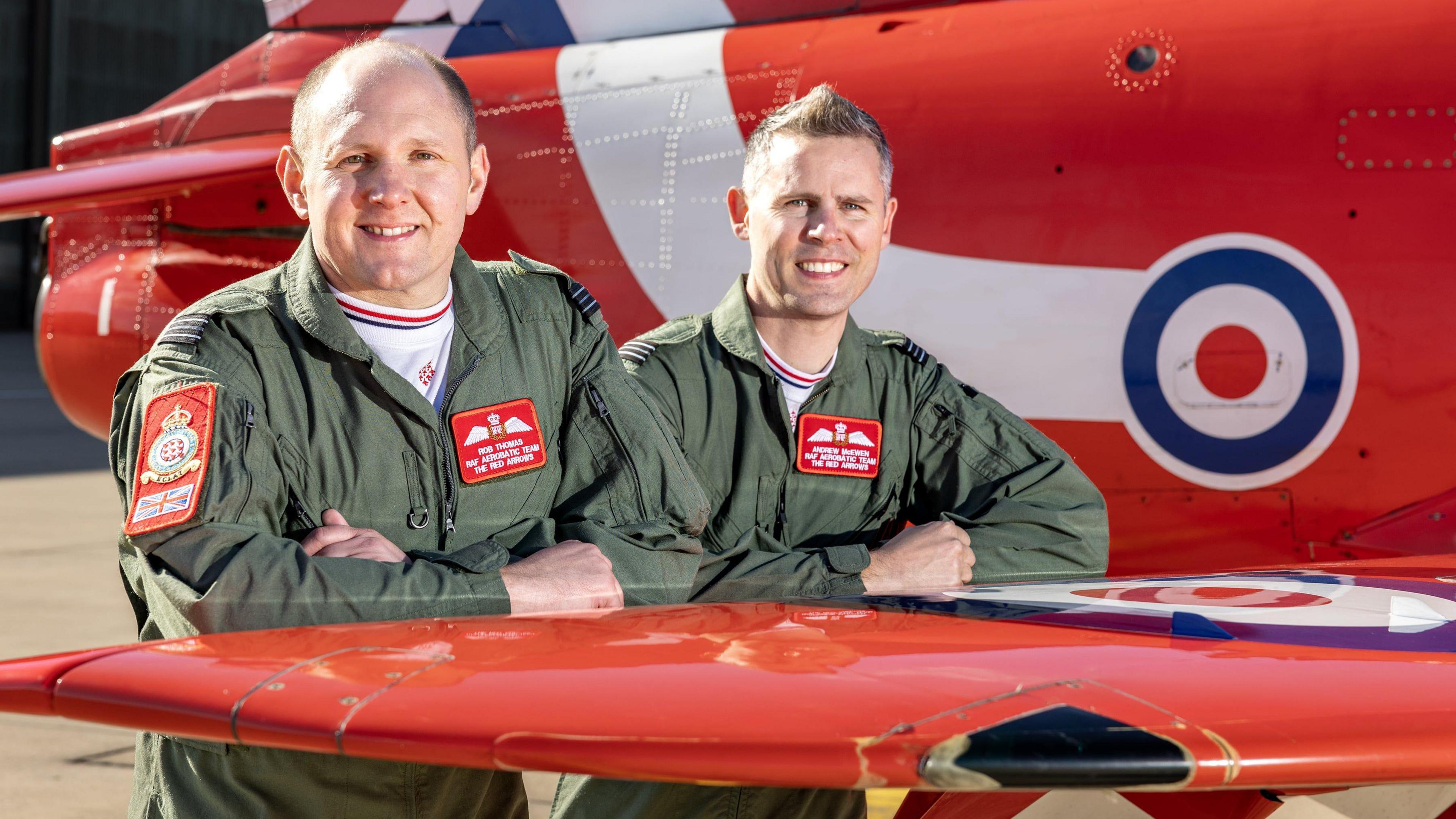 Two men wearing green military clothing, featuring the Red Arrows insignia, pose for a photo on the wing of a Red Arrows plane. They are smiling at the camera.