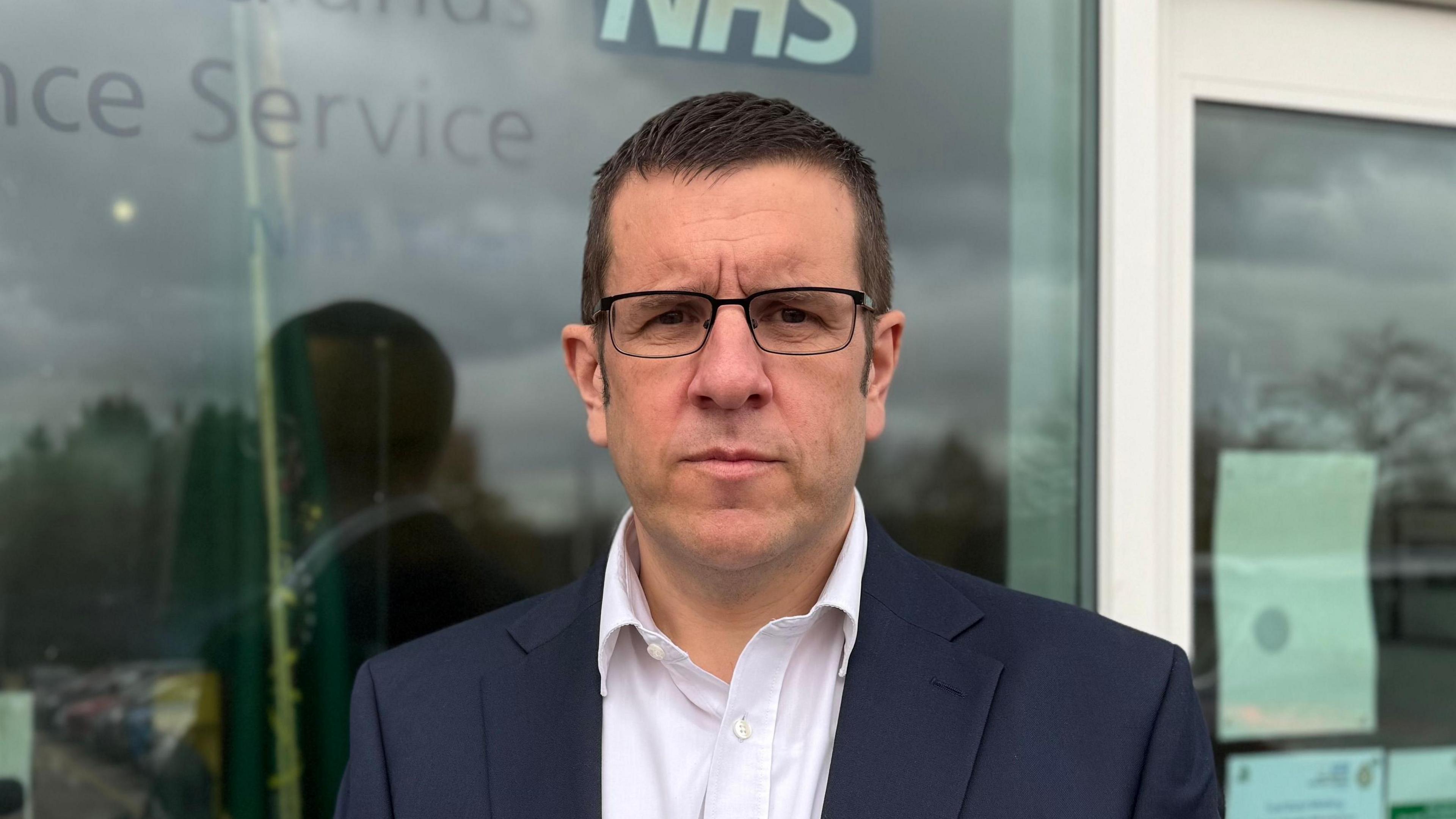 A photograph of a man wearing a suit and open neck shirt while wearing black glasses