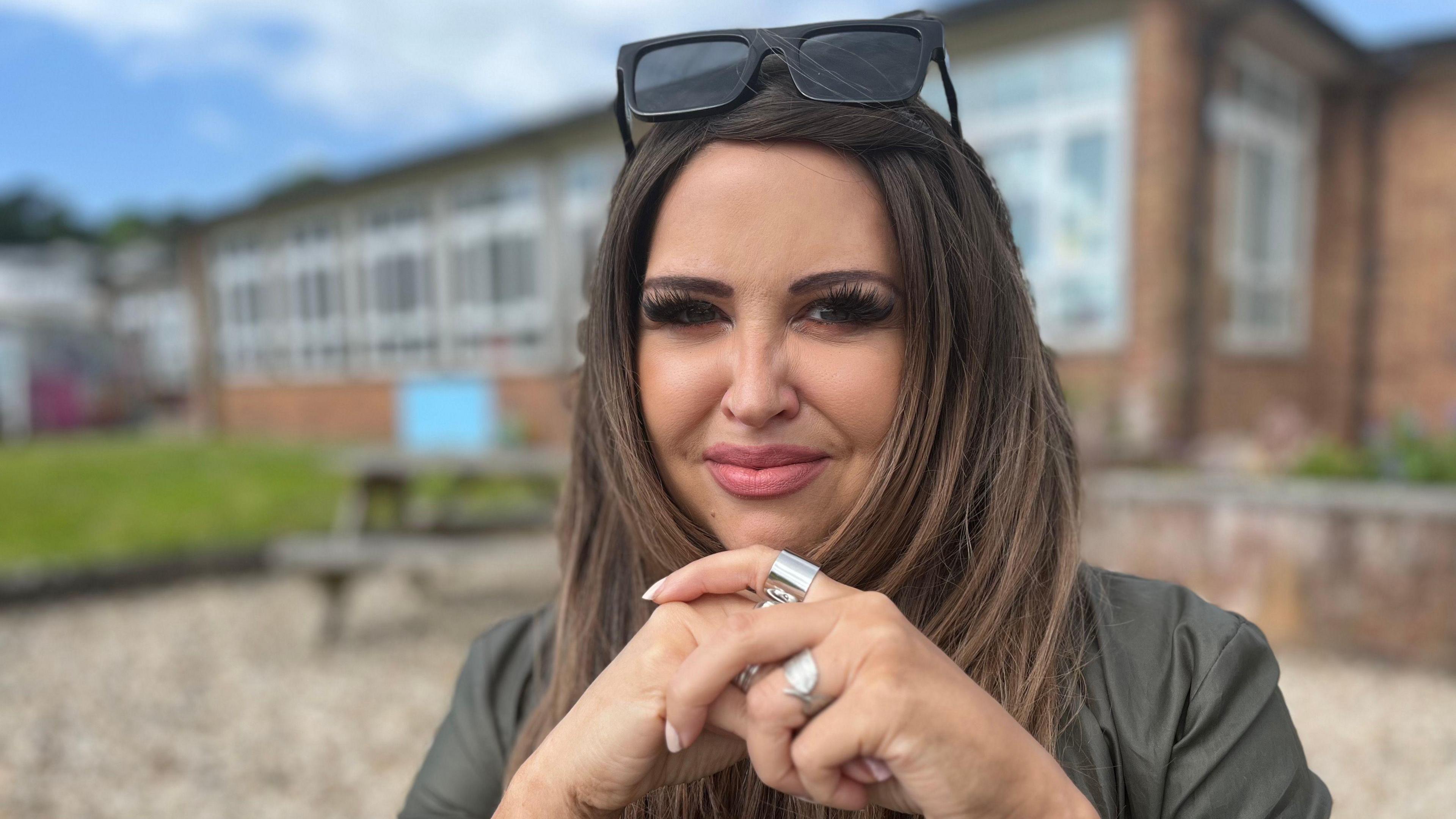 Mared looking at the camera. She is holding her hands up towards her chin and has a couple of silver rings on. She has long dark hair and glasses on her forehead. Behind her is a blurry image of the school.