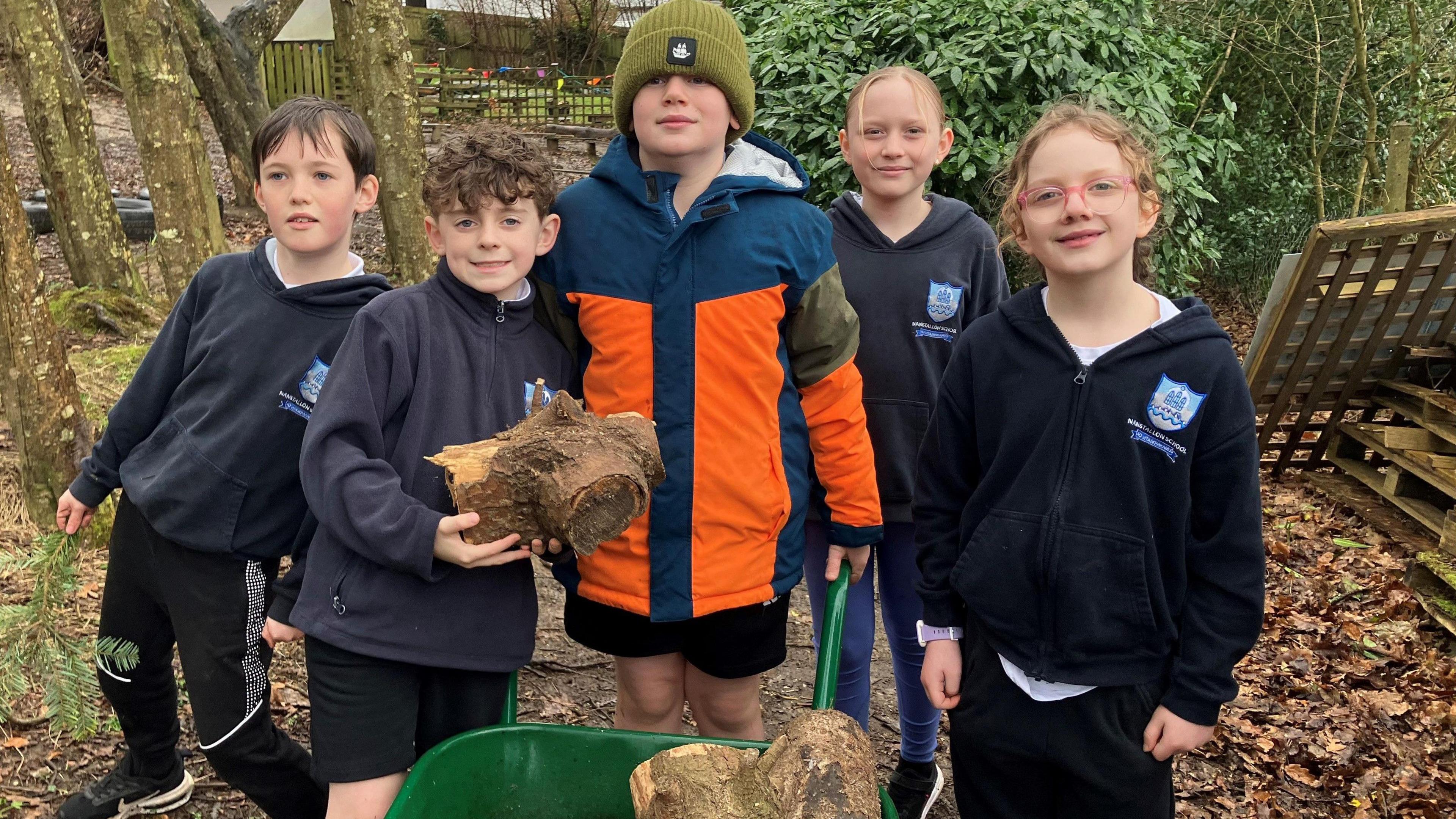 Five children dressed in school uniform in a wooded area using a wheelbarrow to carry wood 