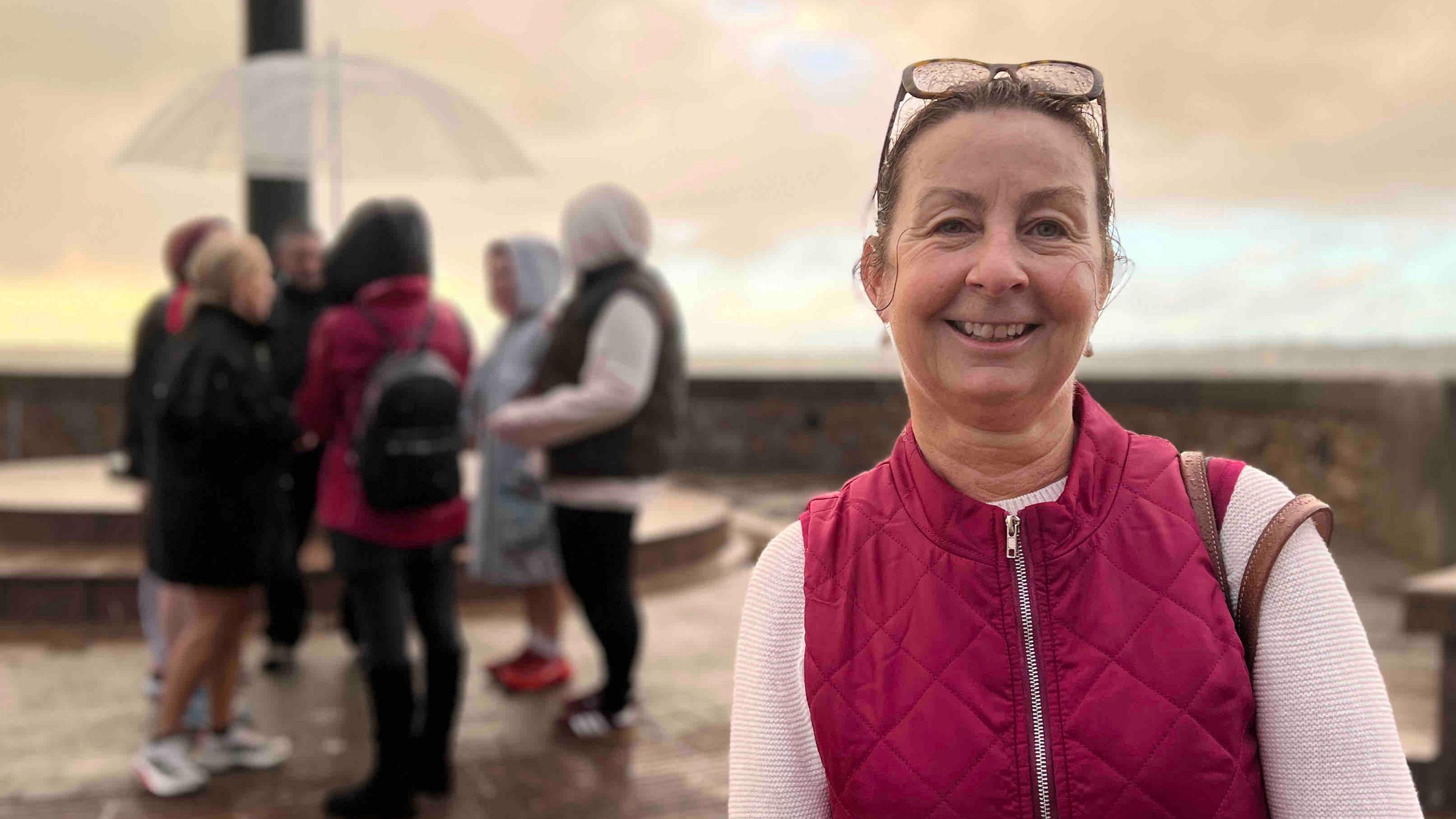 Tracey is wearing a maroon quilted vest over a white long-sleeve top and stands in the foreground. Sunglasses rest on her head, and a teal bag is visible on her left shoulder. In the background, a group of people dressed in casual outdoor clothing gather near a circular stone structure. One person holds a transparent umbrella. The setting appears to be an elevated viewpoint with cloudy skies and wet pavement, suggesting recent rain.