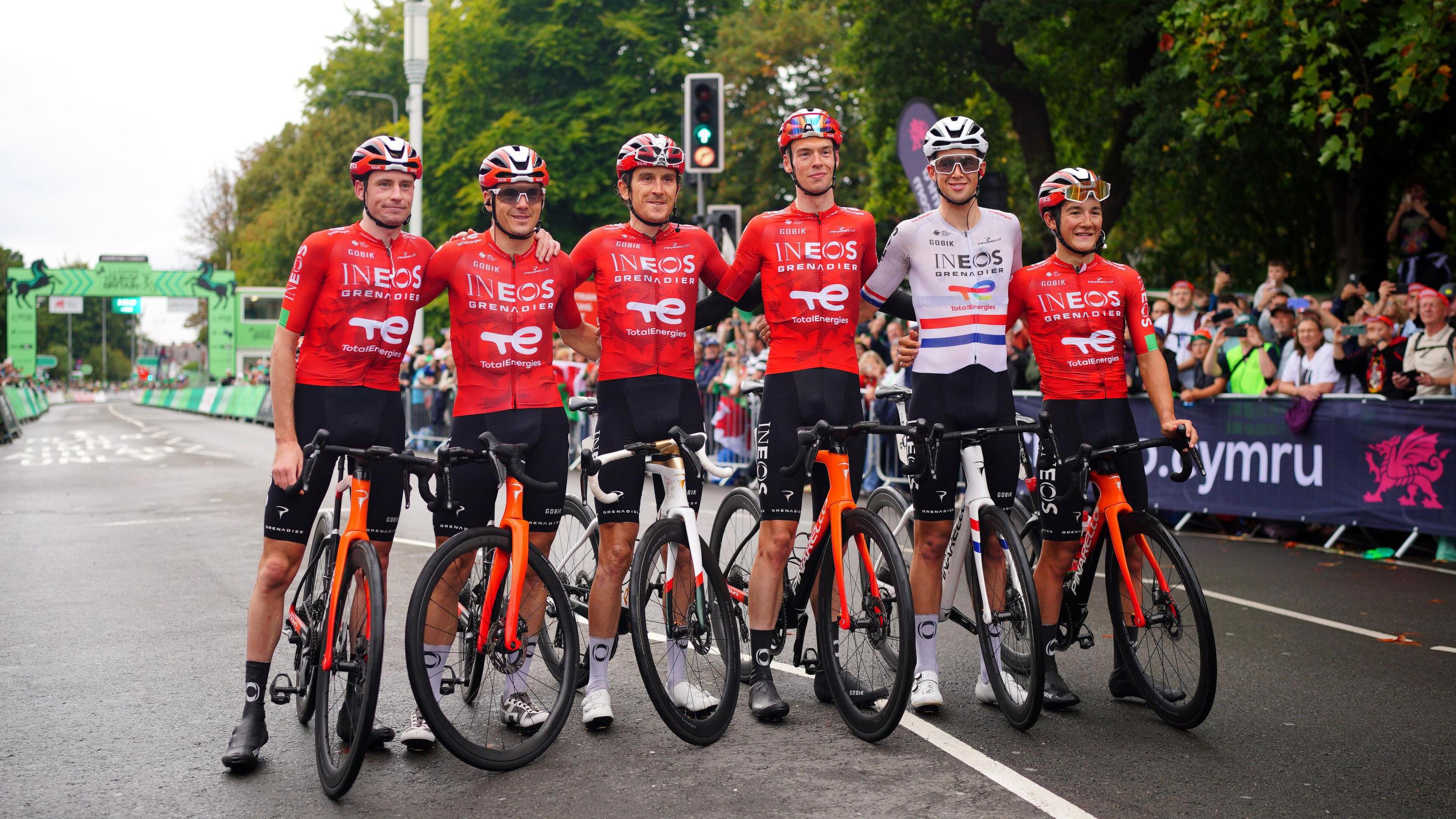 Six men line up to take a photo, five are wearing red jerseys with the one second right wearing a white one. They are all on bikes. Geraint Thomas is third from left. 