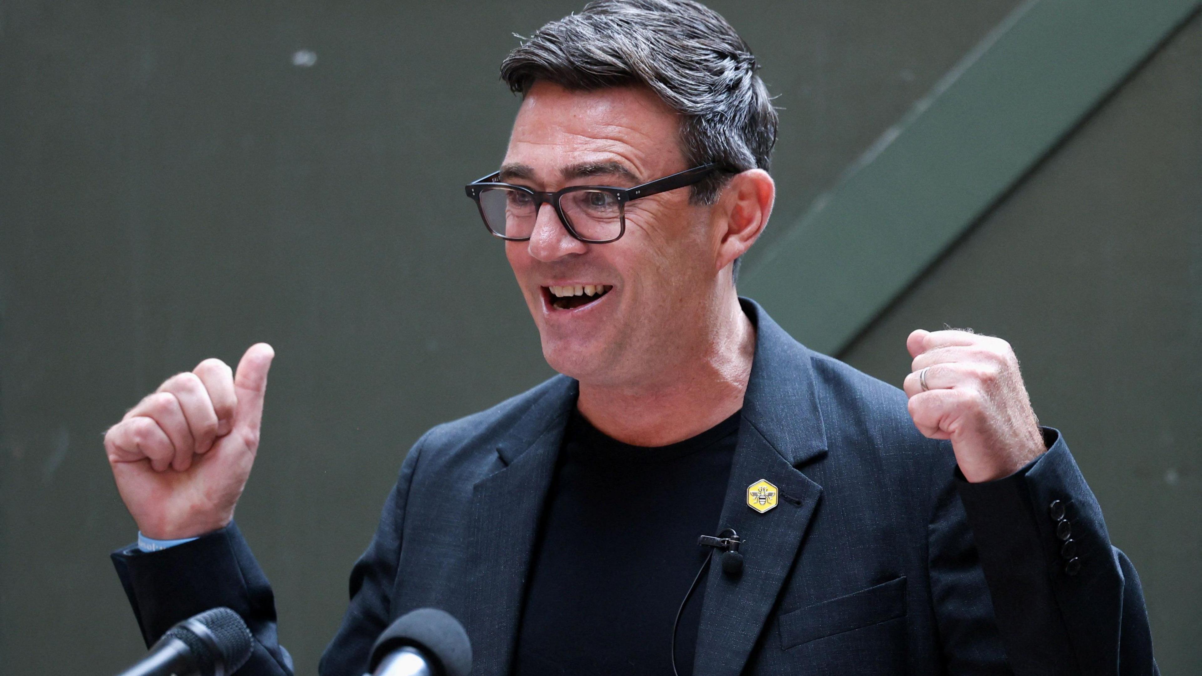 Andy Burnham, wearing a black jacket and t-shirt, stands at a podium.