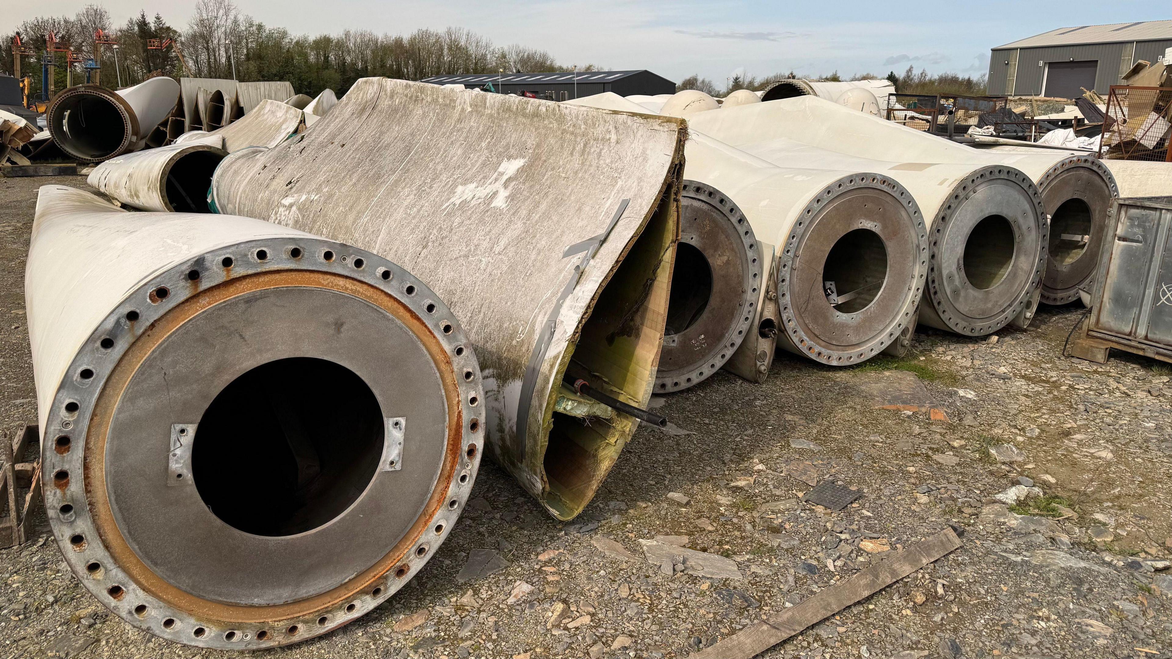 A row of disused wind turbine blades lie in an industrial yard.  The white blades are rounded and hollow at one end but taper off into a flat blade in the distance. There is a warehouse and trees in the distance.