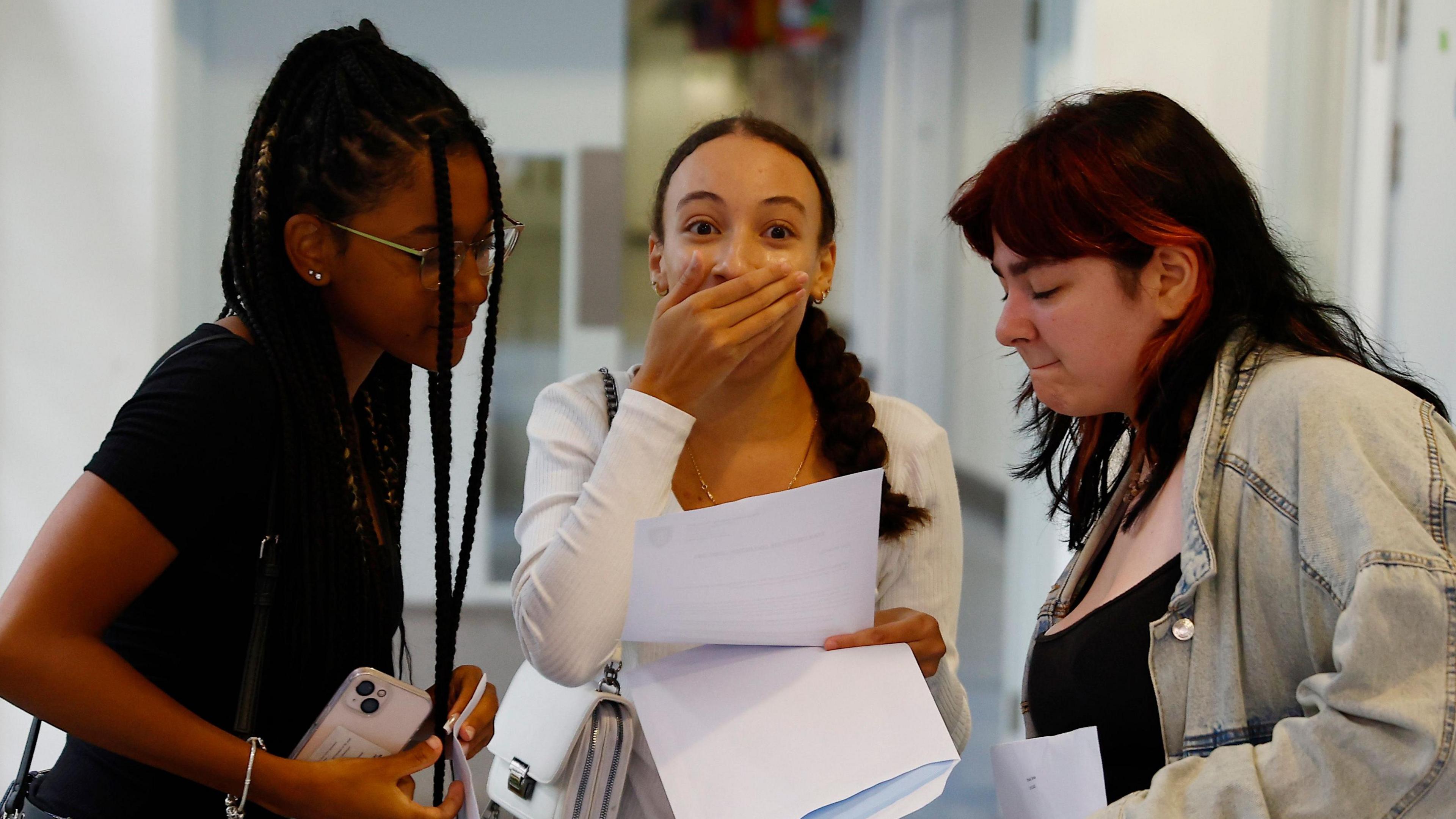 Three teenagers collect their exam results, with one holding their hand over their mouth in shock, and the other two looking at the results